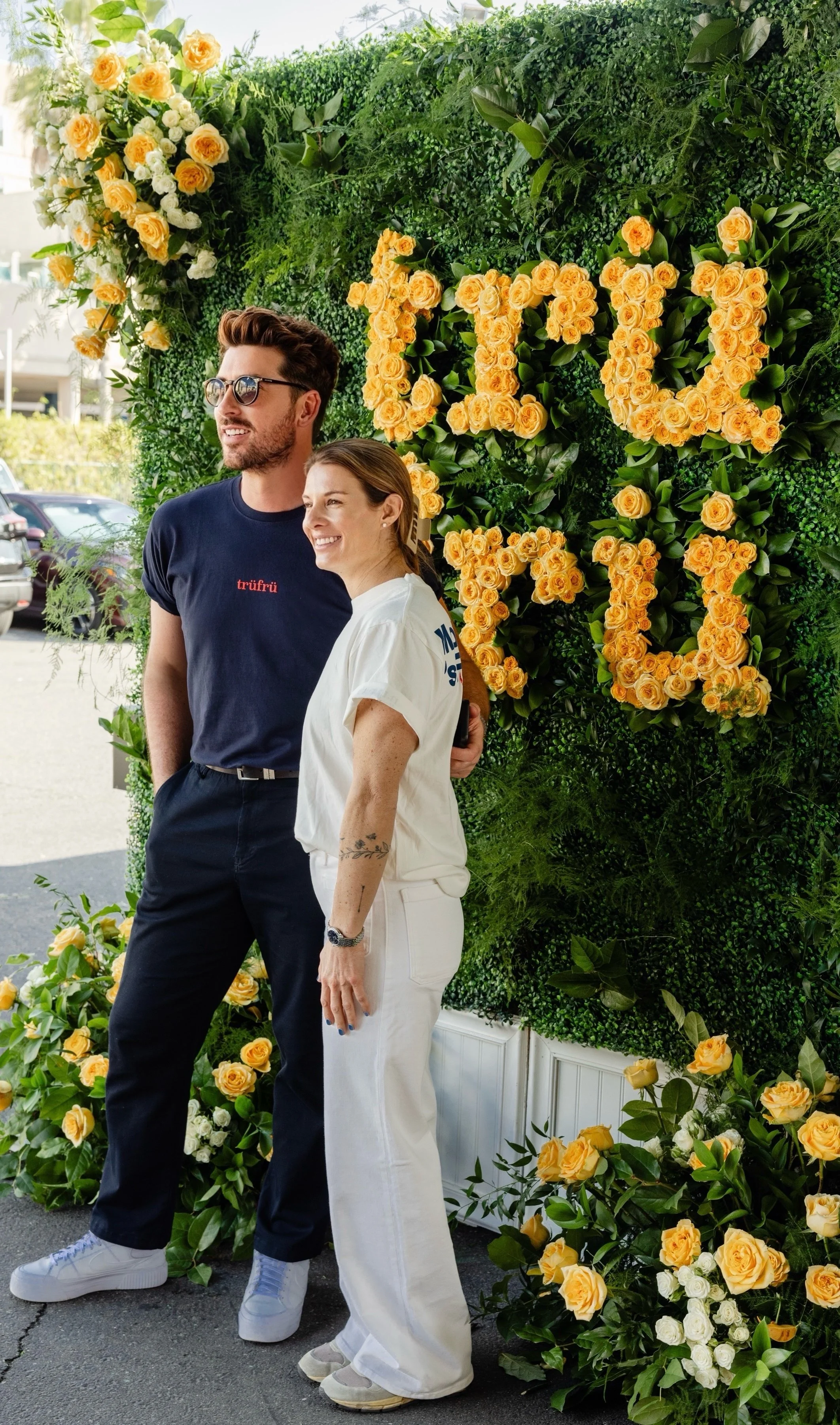 Two people, a man and a woman, standing together outdoors in front of a green floral wall with the words "FLOWER SHOP" spelled out in yellow roses. The man is wearing sunglasses and a navy shirt, while the woman is smiling and dressed in white.