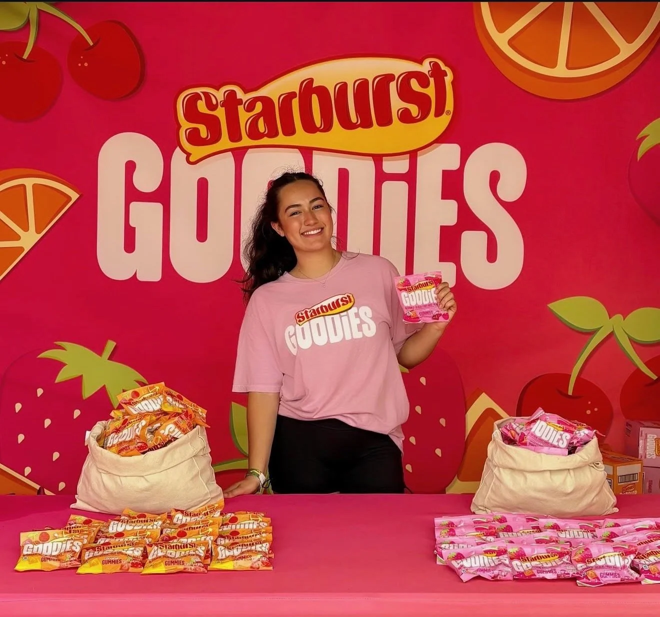 Young woman standing behind a pink table, holding a bag of Starburst Gummies. The background features a pink wall with large Starburst Gummies branding and illustrations of cherries and orange slices. Several bags of Starburst Gummies are displayed on the table.