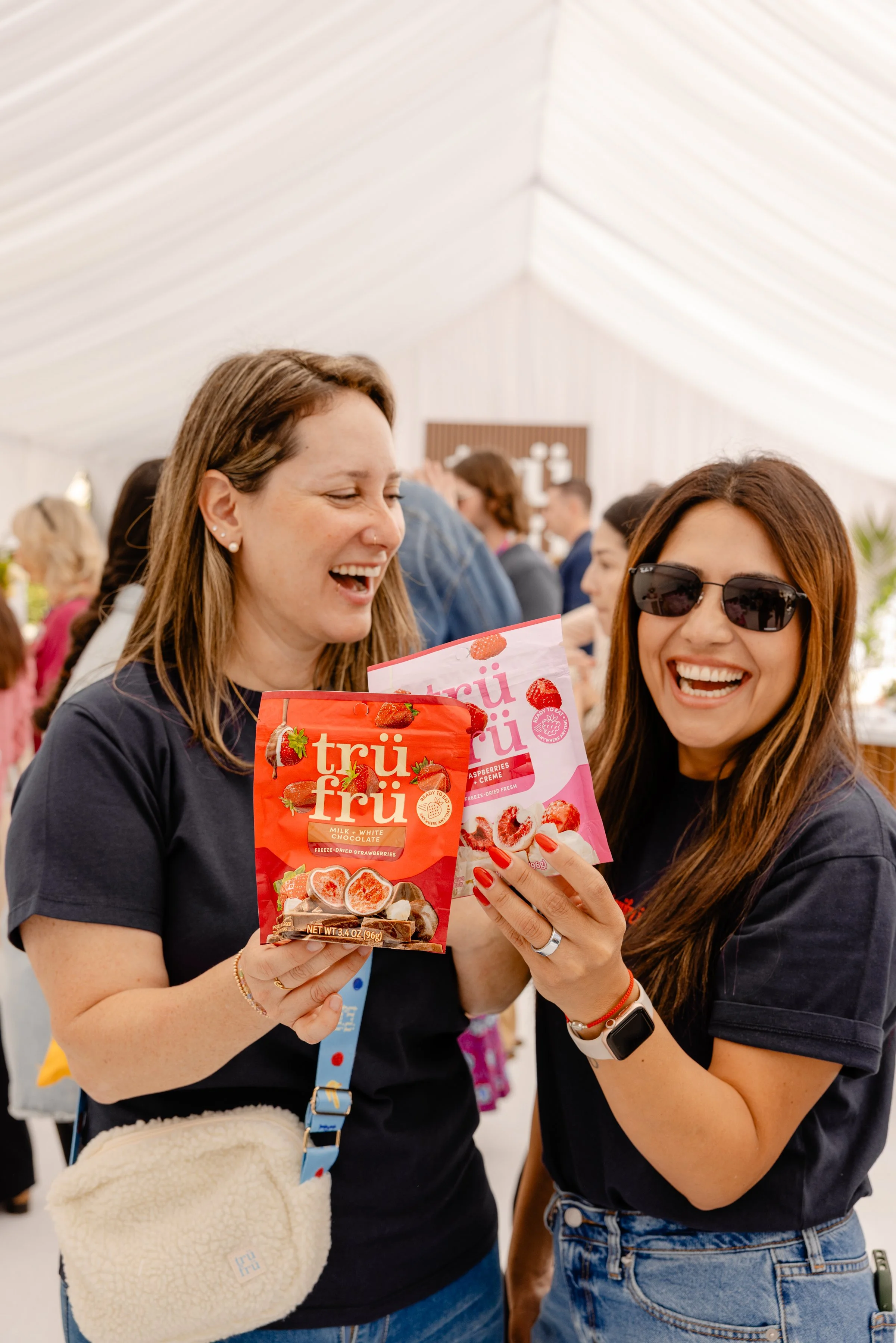 Two women laughing and holding packages of Trüf fri freeze-dried strawberries at an indoor event.