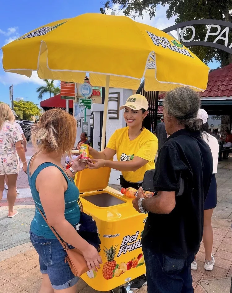 Del Frutal brand ambassador handing a juice sample to a shopper from a yellow branded cart