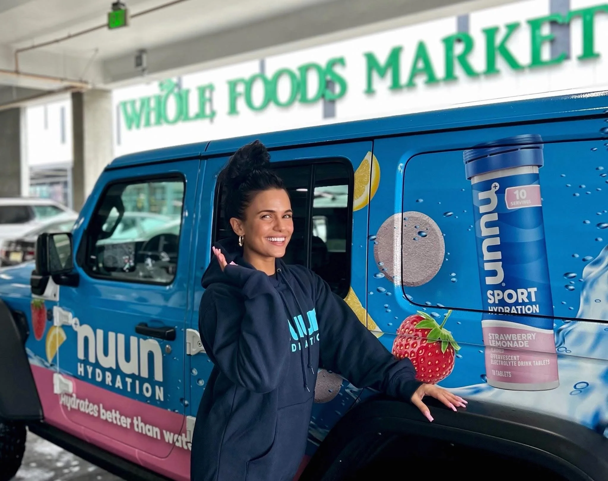 Woman smiling and pointing at a blue hydration drink truck parked outside Whole Foods Market.