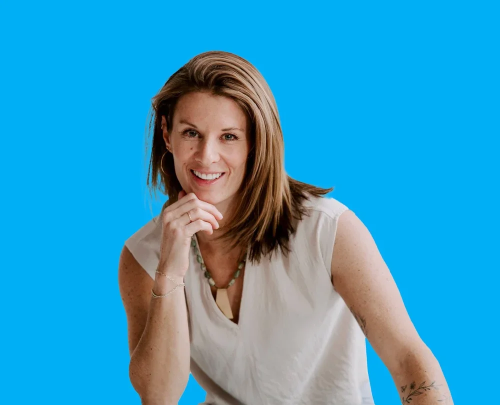 A smiling woman with straight, shoulder-length brown hair, wearing a sleeveless white top, gold hoop earrings, a beaded necklace, and a tattoo on her left arm, posing in front of a bright blue background.