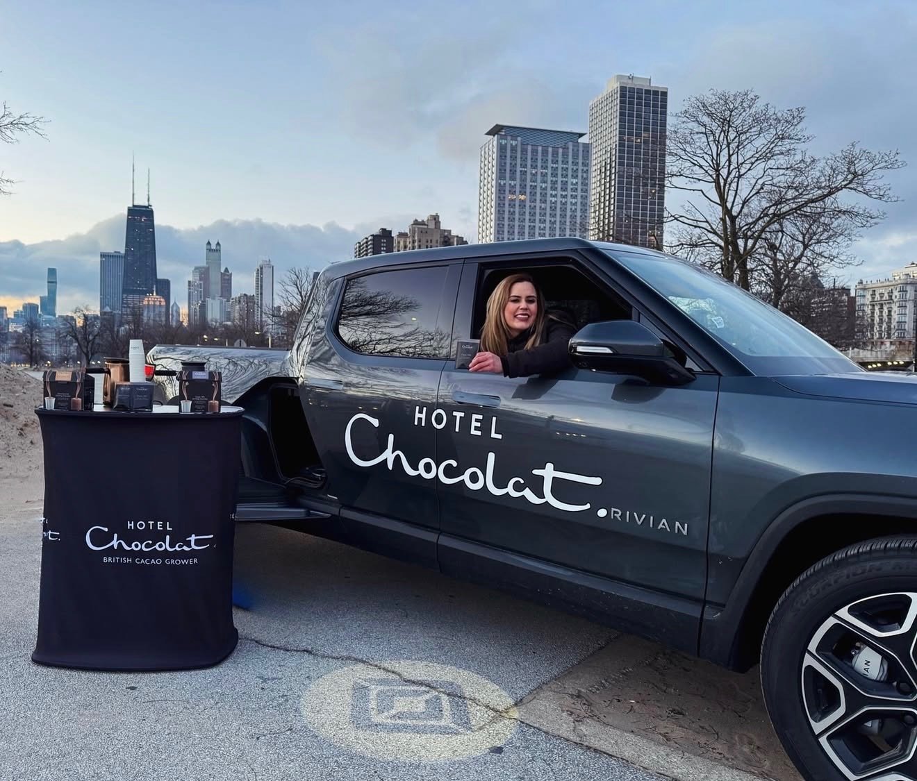 A woman sitting in a black SUV with 'Hotel Chocolat' branding, parked outdoors in a city area with tall buildings and a cloudy sky in the background, next to a display table with chocolate products.