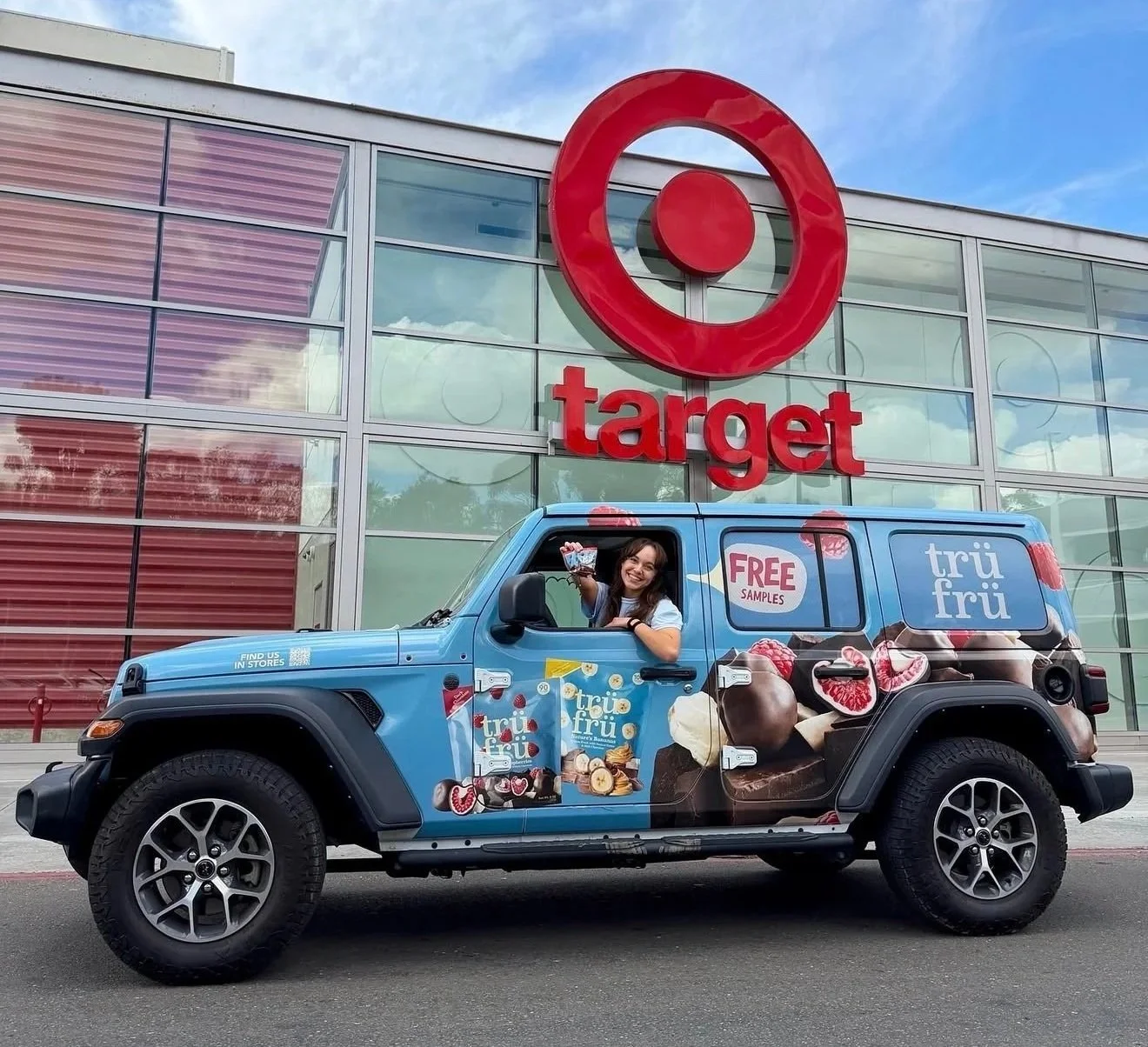 A woman sitting in a Jeep with Tru Frū branding, parked in front of a Target store, holding a water bottle and smiling, with a promotional sign for free samples on the vehicle.