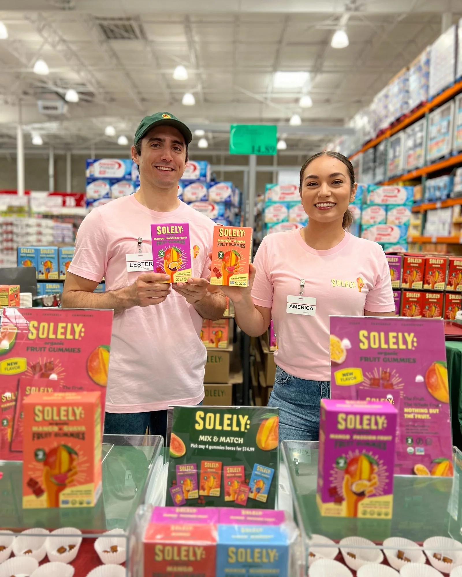 Two brand ambassadors in pink SOLELY tees holding boxes of SOLELY organic fruit gummies at a Costco roadshow sampling table