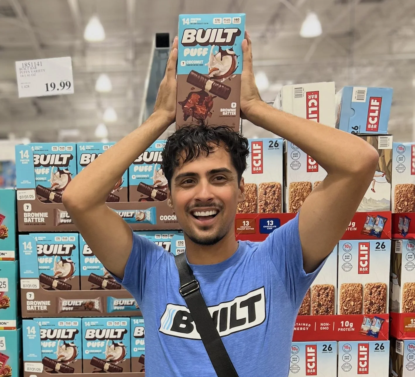 Smiling brand ambassador in a blue BUILT tee holding a box of BUILT Puff coconut brownie batter bars on top of his head in front of a Costco wall of BUILT Puff boxes and a price sign