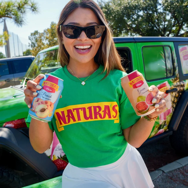 Natura's brand ambassador in a green Natura's tee holding peach and guava nectar cans beside a Natura's-wrapped Jeep