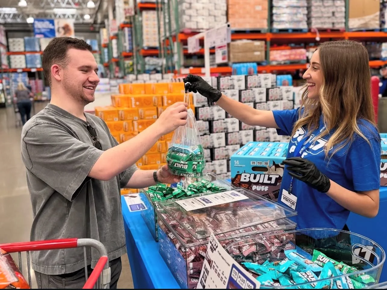Front & Center brand ambassador in a BUILT tee and black gloves handing a bag of BUILT bar samples to a smiling Costco member with a cart at a Costco roadshow