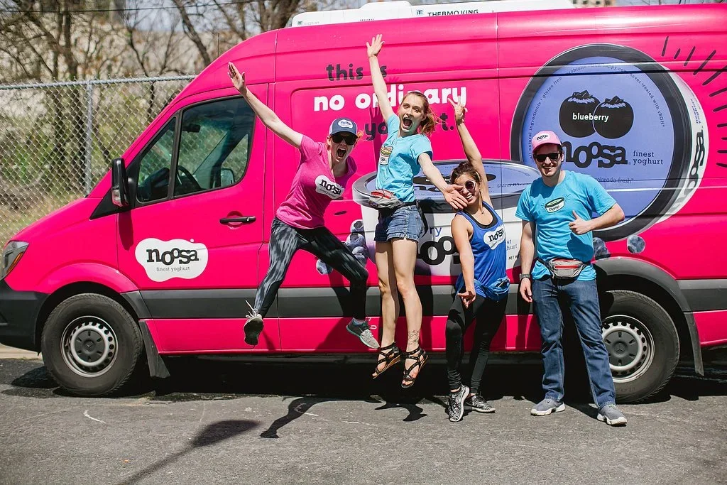 Four people posing excitedly in front of a large, bright pink yogurt truck with Nosa branding and a blueberry yogurt image, outdoors on a sunny day.