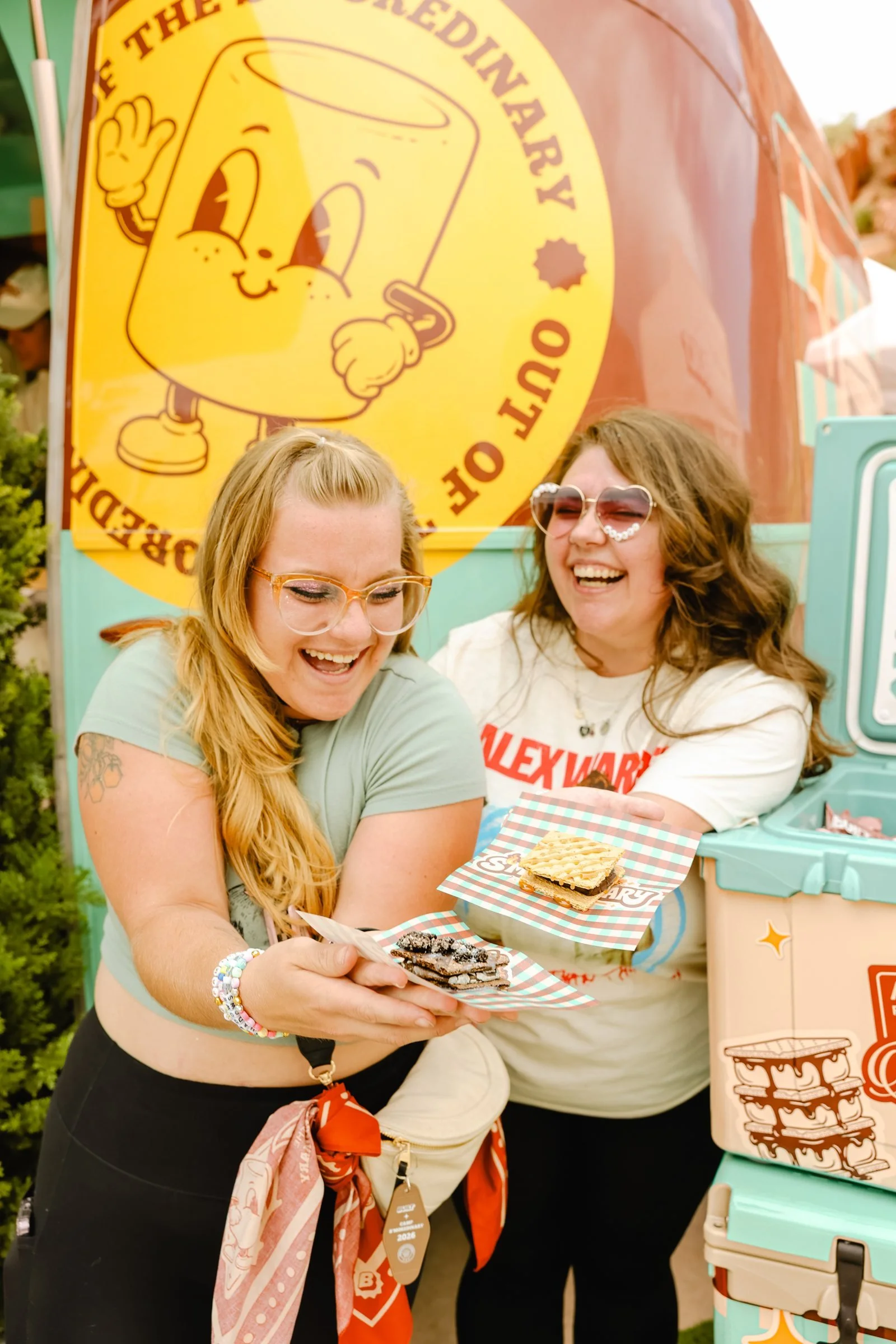 Two friends laughing as they hold up s&rsquo;mores in front of the Camp S&rsquo;moredinary camper