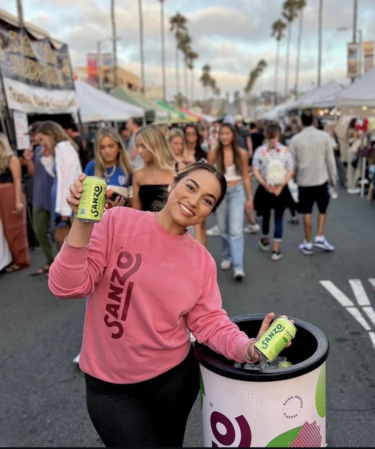 Ambassador in a pink Sanzo sweatshirt sampling cans at a busy street festival