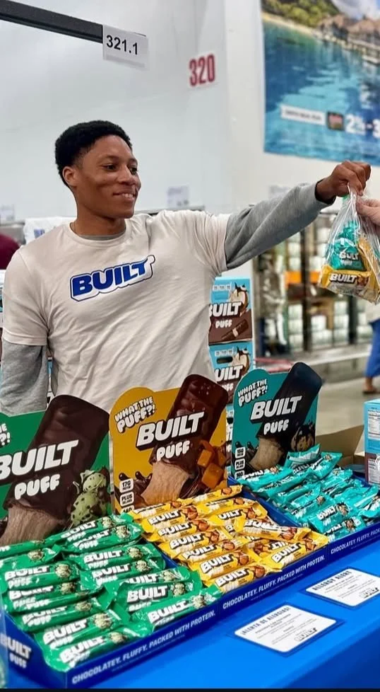 Brand ambassador handing a BUILT Puff sample bag across a Costco roadshow display table