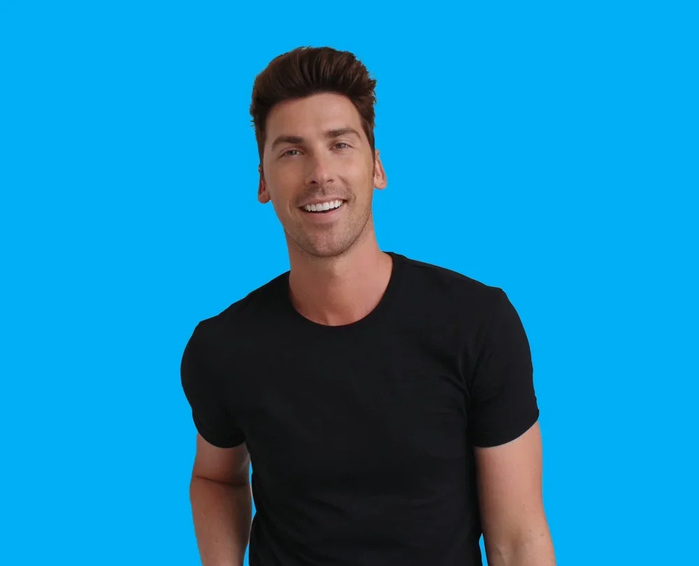 A man with brown hair, wearing a black t-shirt, smiling against a bright blue background.