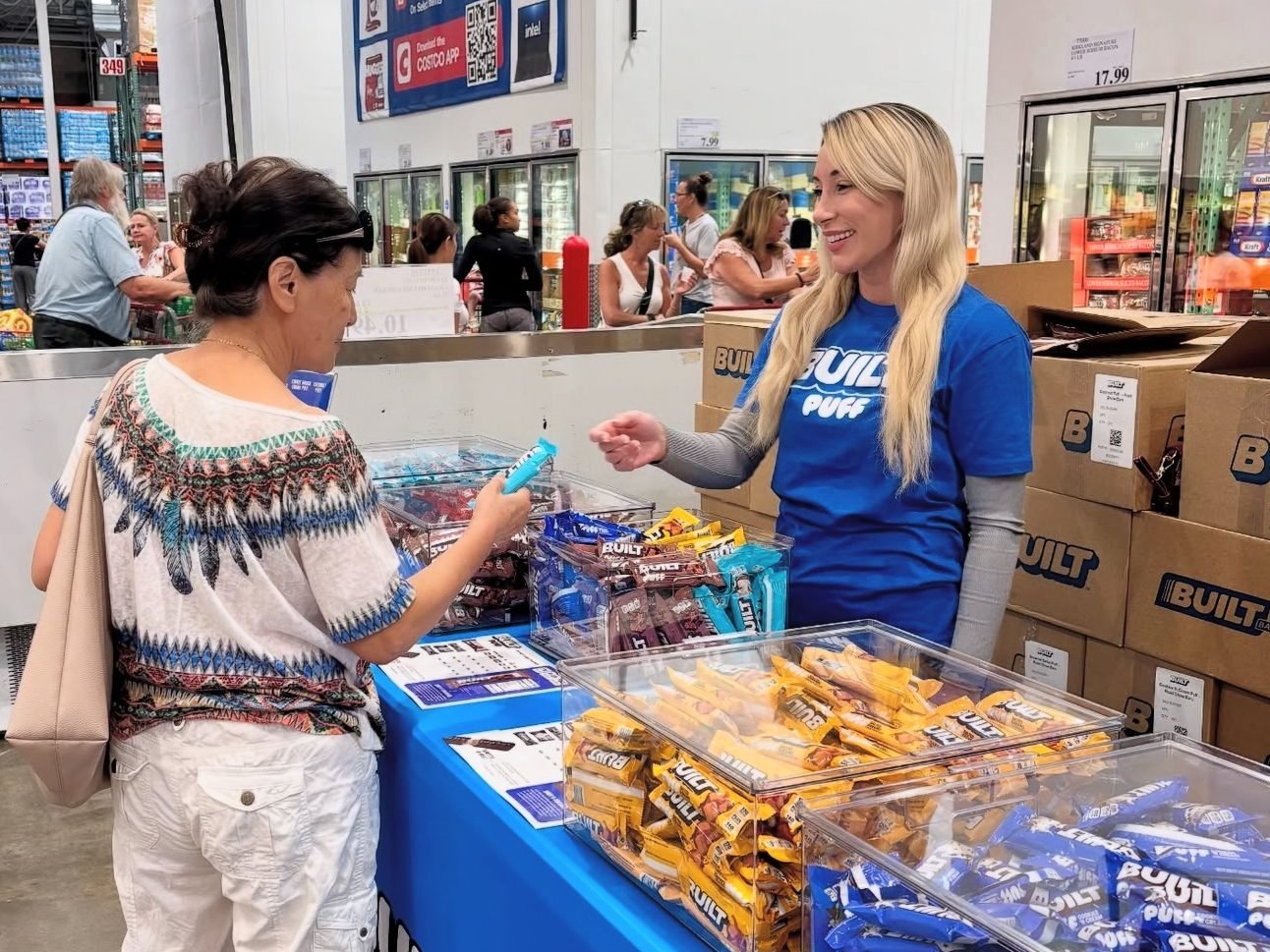 Brand ambassador in a BUILT tee offering a BUILT bar sample to an older woman and talking her through it at an in-warehouse Costco demo table