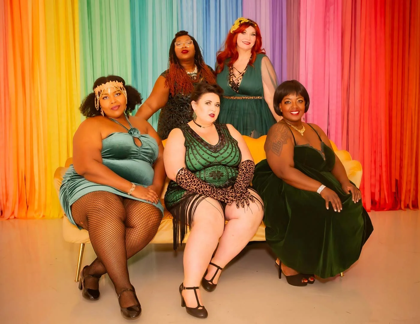 Six women from Down South Delectables Burlesque Collective in colorful dresses sitting and standing in front of a rainbow-colored curtain, posing for a photo.