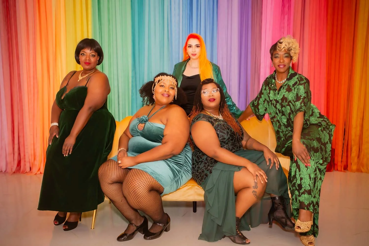 A group of six women from Down South Delectables Burlesque Collective in colorful dresses posed in front of a rainbow-colored curtain background.