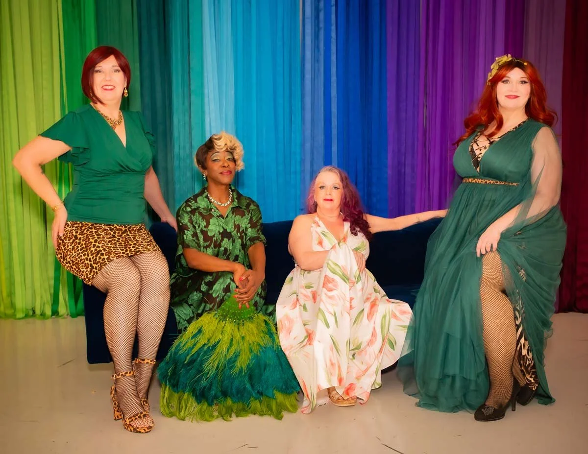 Five women from Down South Delectables Burlesque Collective in colorful dresses sitting and standing in front of a rainbow-colored curtain.