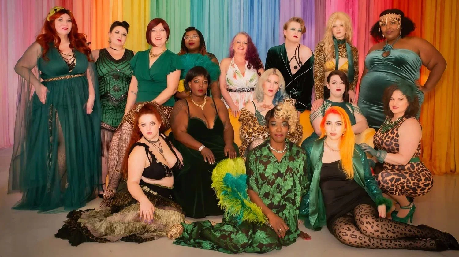 Group of women from Down South Delectables Burlesque Collective dressed in colorful, vintage-inspired clothing posing in front of a rainbow-colored curtain.