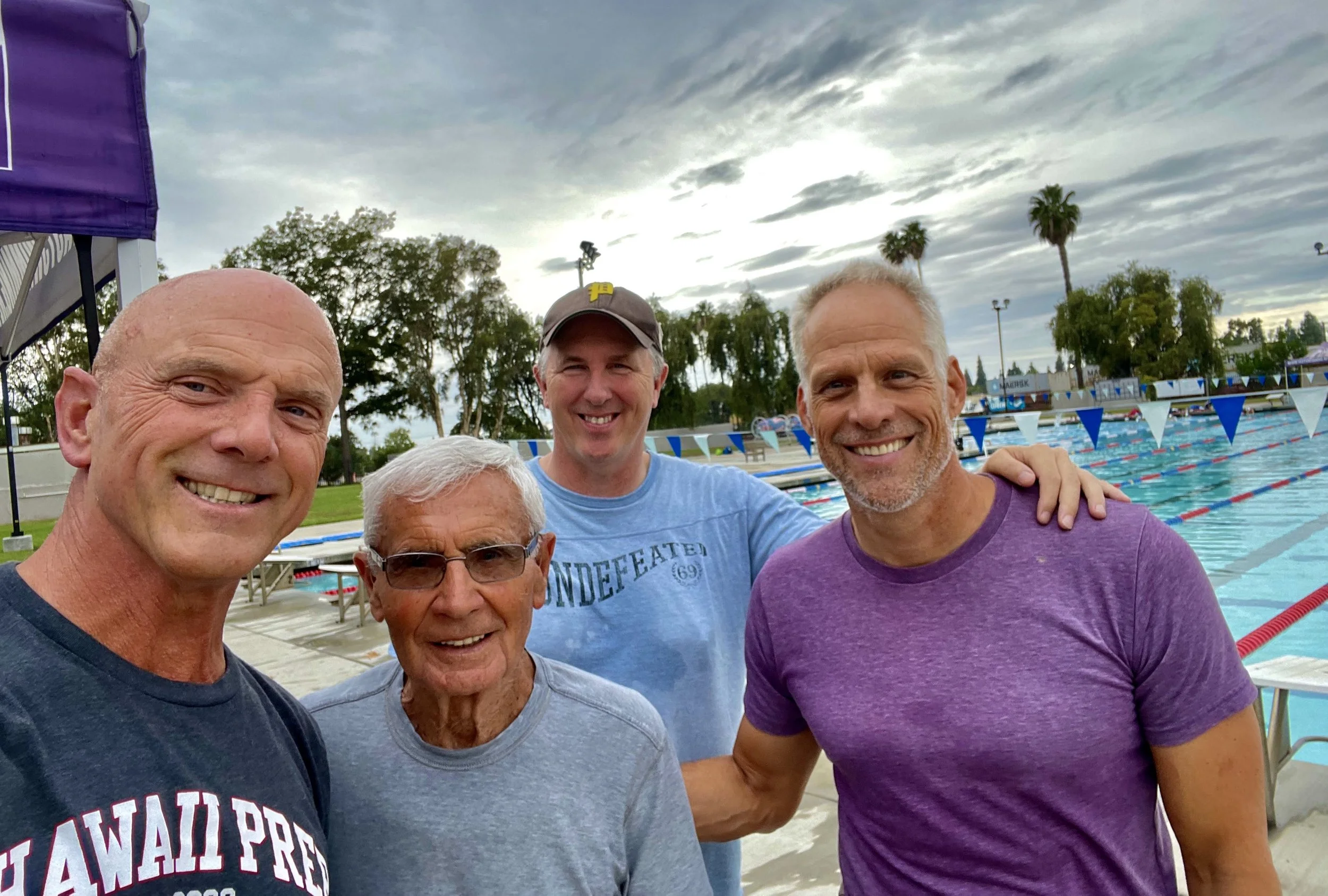 Five men standing together at an outdoor swimming pool, smiling. The pool has swimming lanes with blue and white lane dividers, and there are trees and cloudy skies in the background.
