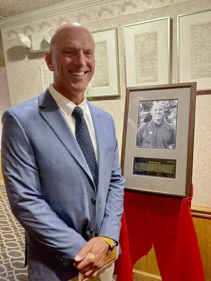 A smiling man in a blue suit with a white shirt and dark polka-dot tie standing next to a framed black-and-white photograph and plaque at an indoor event.