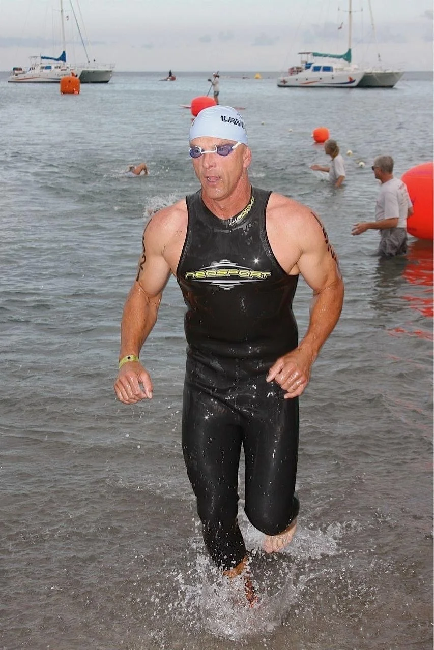 A man in a wetsuit emerging from the water at a beach, wearing goggles and a swim cap, with boats in the background and other people swimming nearby.