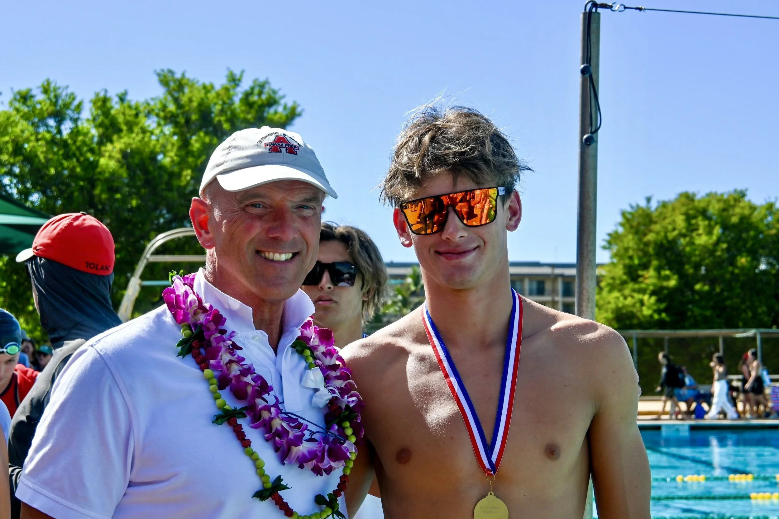 An older man wearing a white cap and leis is standing next to a young man with a medal around his neck, sunglasses, and no shirt at an outdoor swimming pool, celebrating after a swim event.