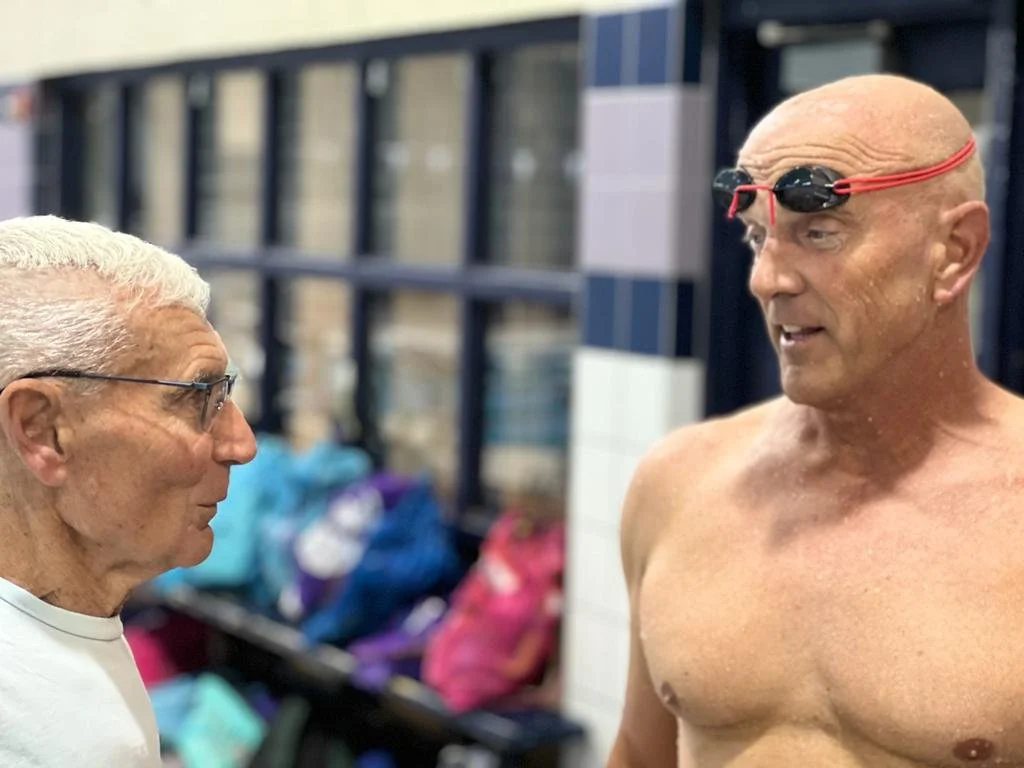 Two men, one older with white hair and glasses, and one younger, shirtless, wearing goggles on his head, talking at an indoor pool.