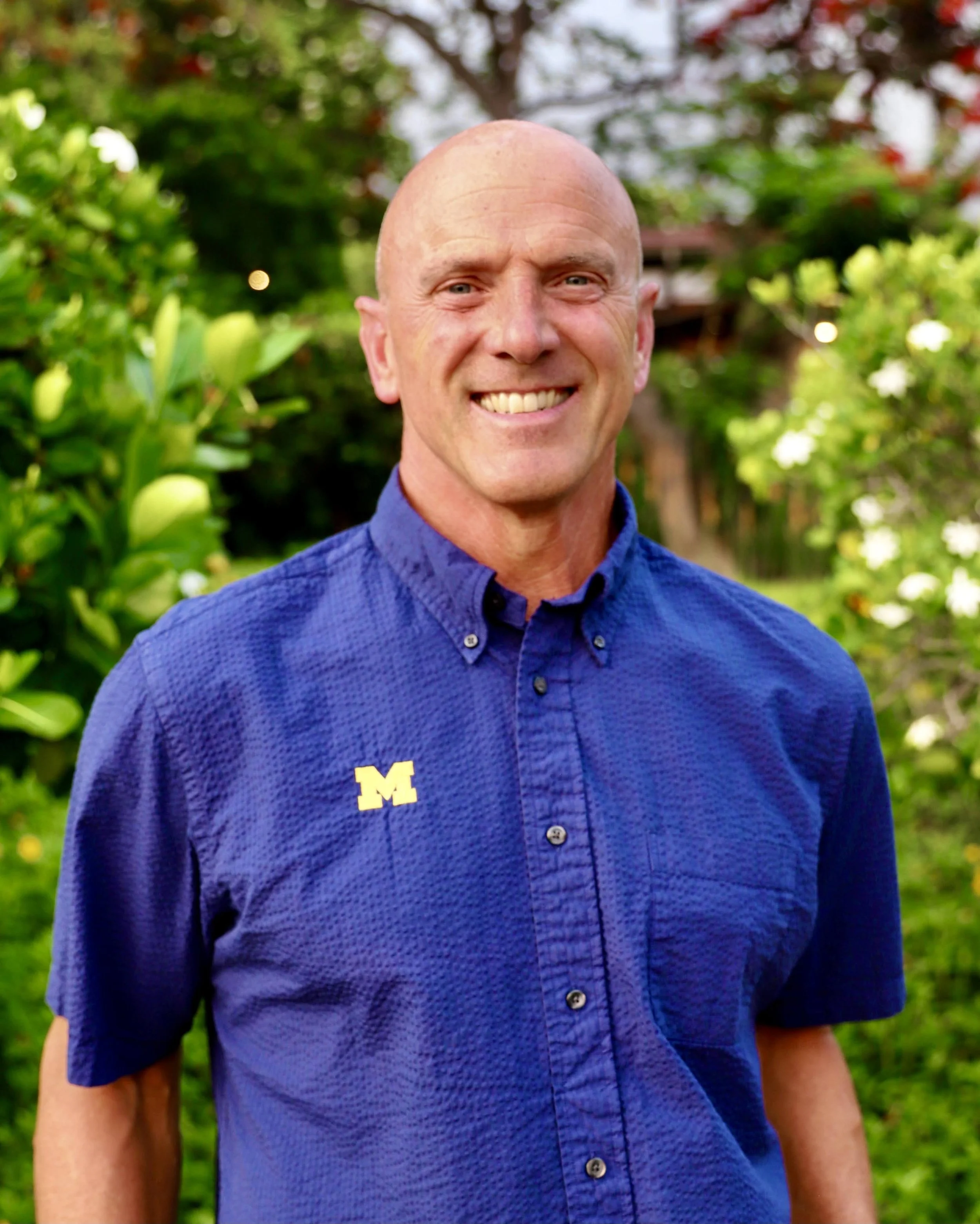 A smiling man with a shaved head wearing a blue button-up shirt with a yellow 'M' logo, standing outdoors in front of green trees and bushes.