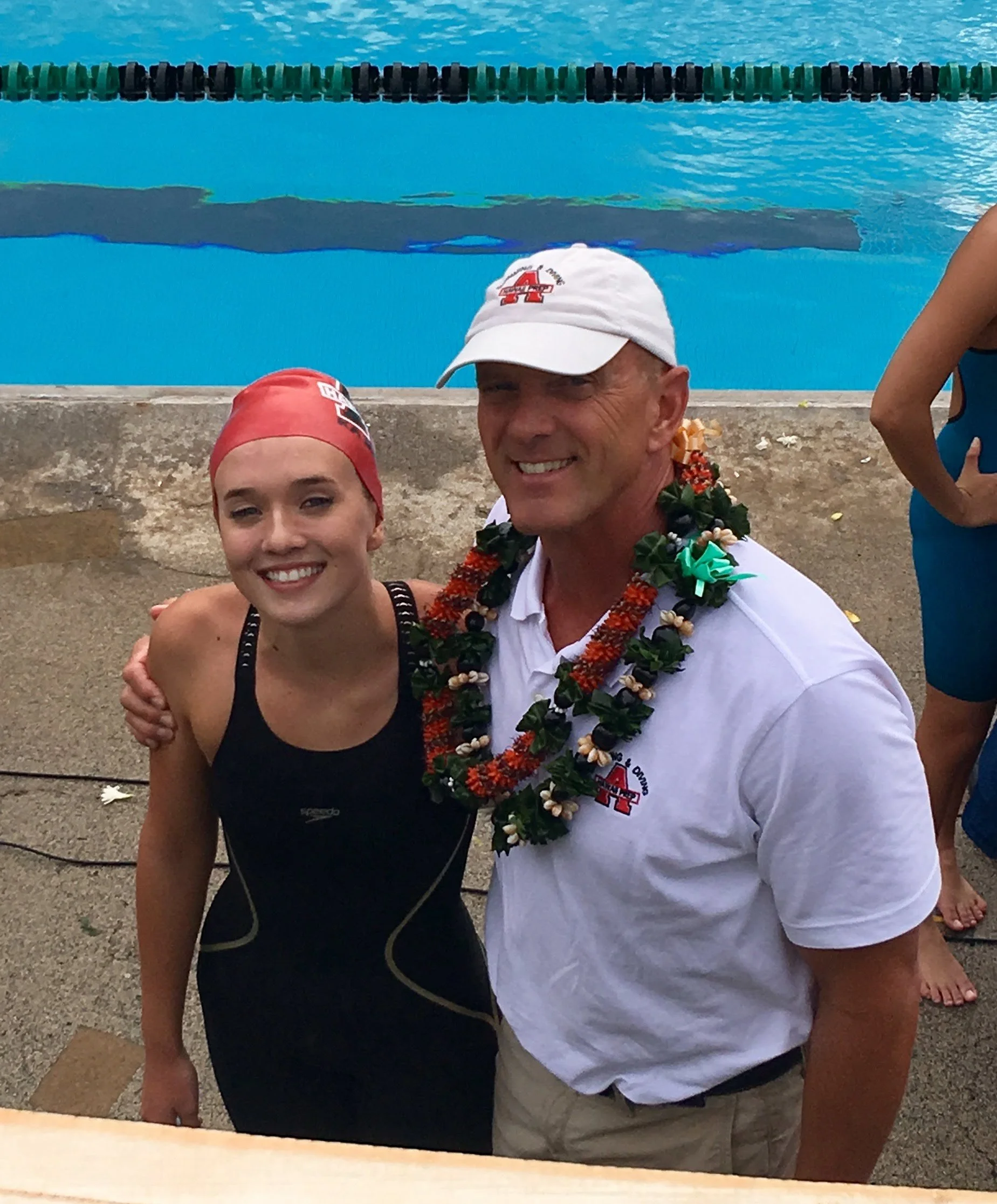 Two people in swimming attire, one man wearing a white cap and a floral lei, and one woman wearing a red swim cap, smiling outdoors near a swimming pool with lane dividers.