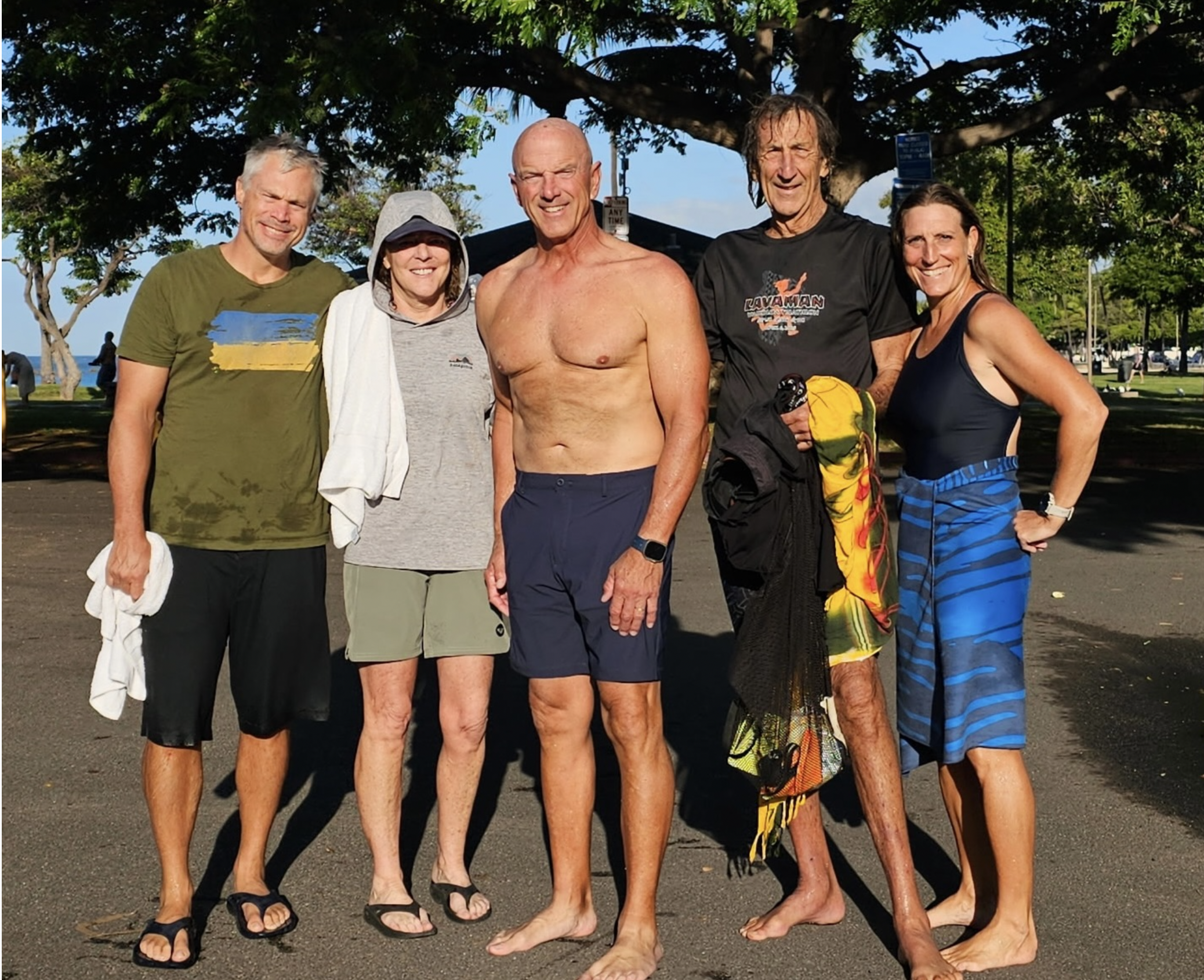 Five friends standing outside under a large tree, smiling, dressed in casual summer attire, with ocean and park background.
