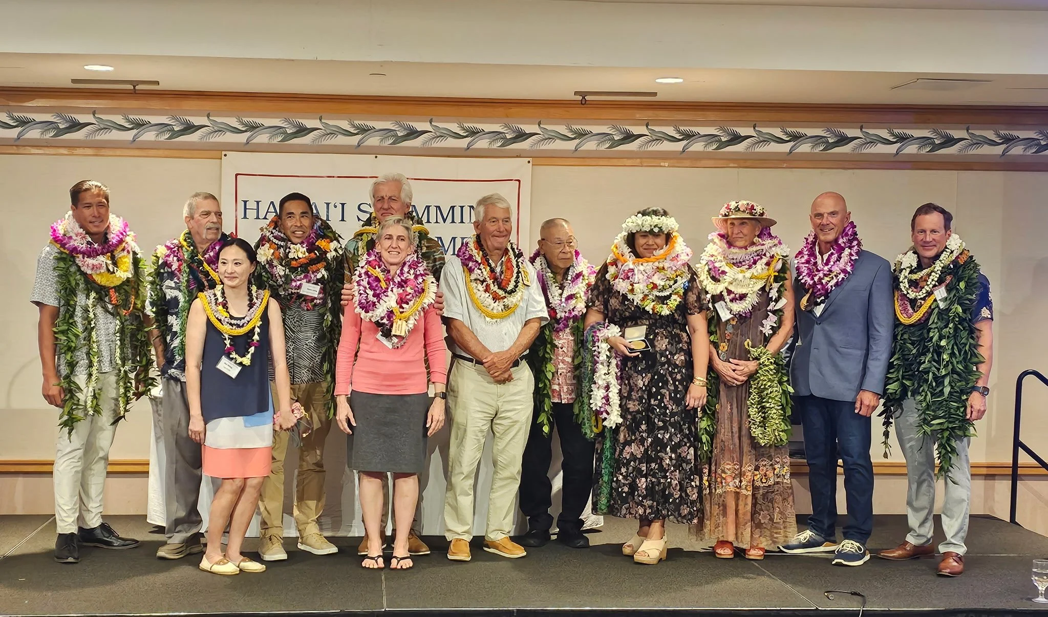 A group of people on a stage at a Hawaii summit, wearing leis and standing in front of a sign that reads "Hawaii Summit." The group includes men and women, some in casual attire and others in formal or traditional Hawaiian clothing. Some individuals 