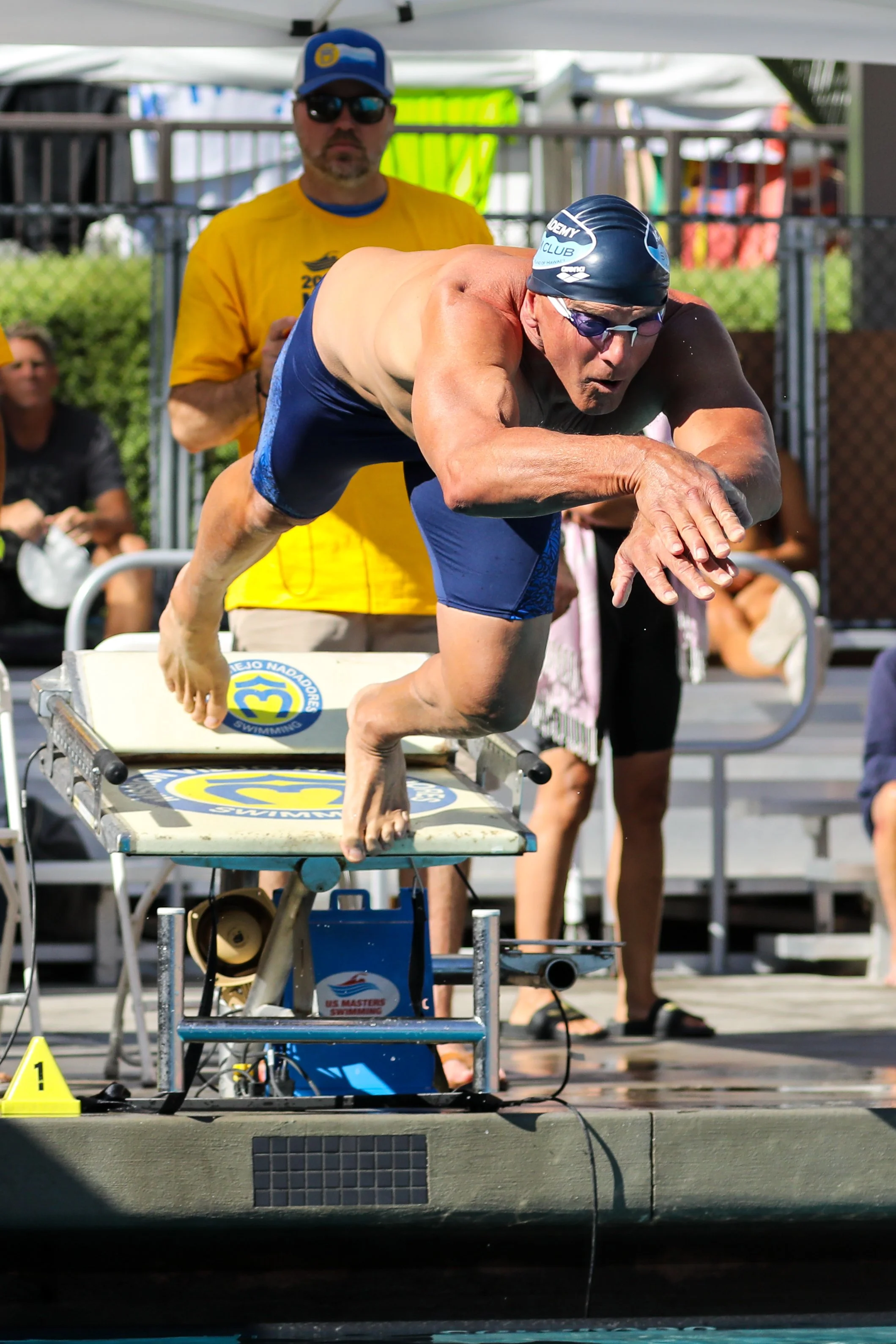 Swimmer in a black swim cap and blue swimwear, crouching on a starting block during a race, with a coach or official in a yellow shirt and sunglasses in the background.