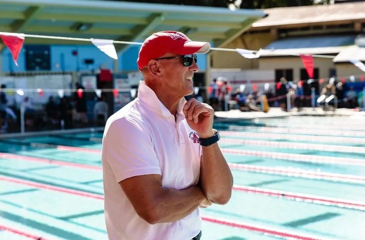 A man in a red cap, sunglasses, and a white polo shirt standing at the edge of a swimming pool, smiling with his arms crossed and hand on his chin, with people and swimming lanes in the background.