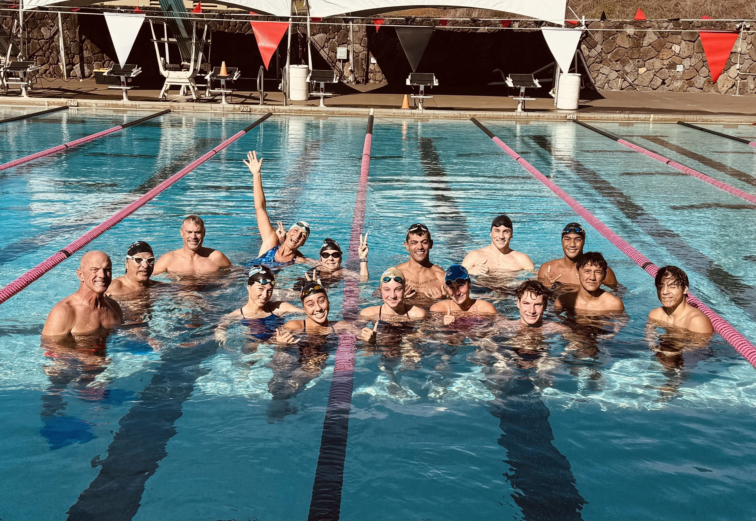 Group of swimmers and coaches celebrating at the end of a swim race in a swimming pool, some making peace signs, with lane dividers and starting blocks visible.