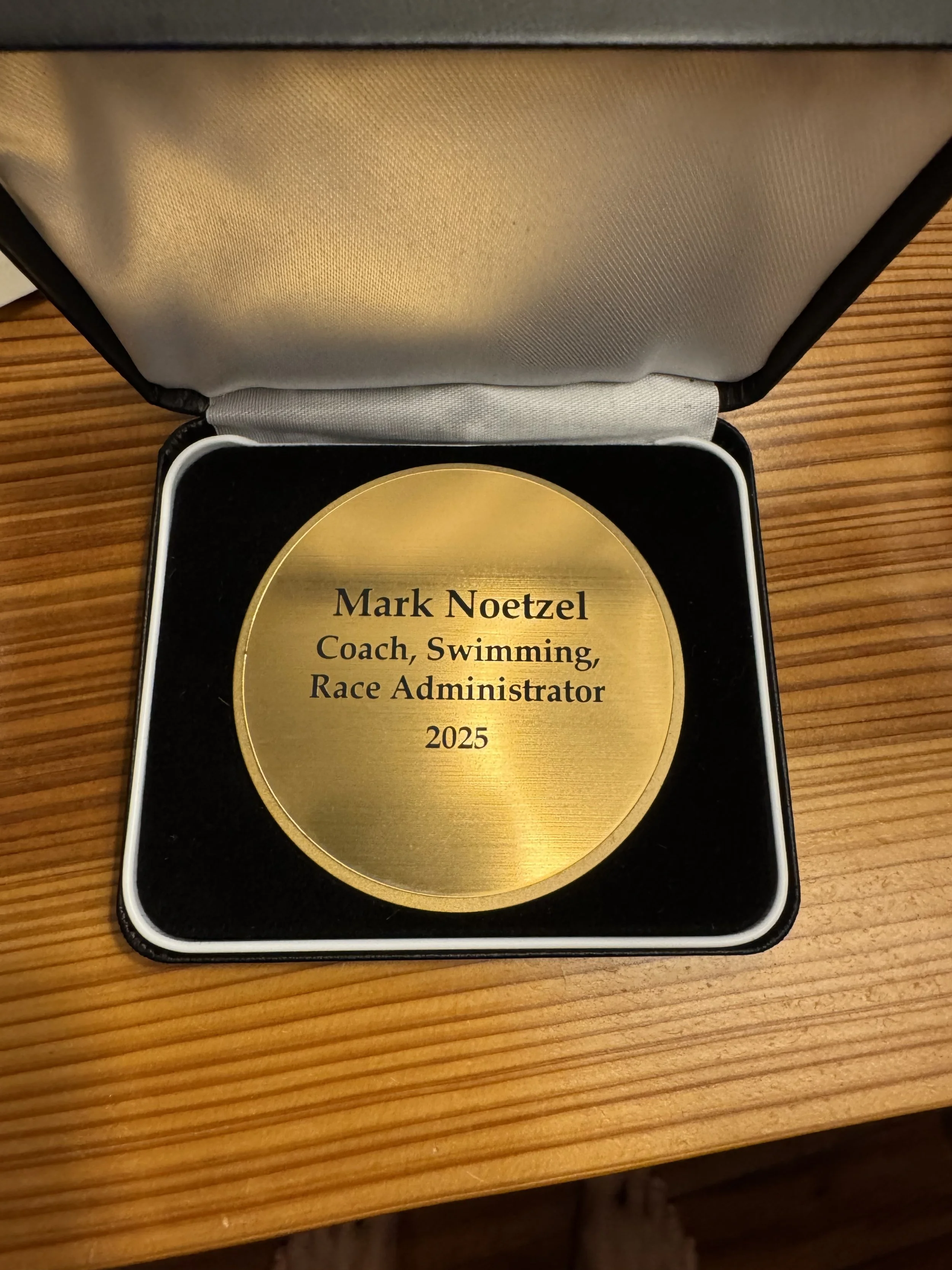 Gold colored medal or plaque with black text that reads "Mark Noetzel Coach, Swimming, Race Administrator 2025" inside a black box with a white trim, placed on a wooden surface.