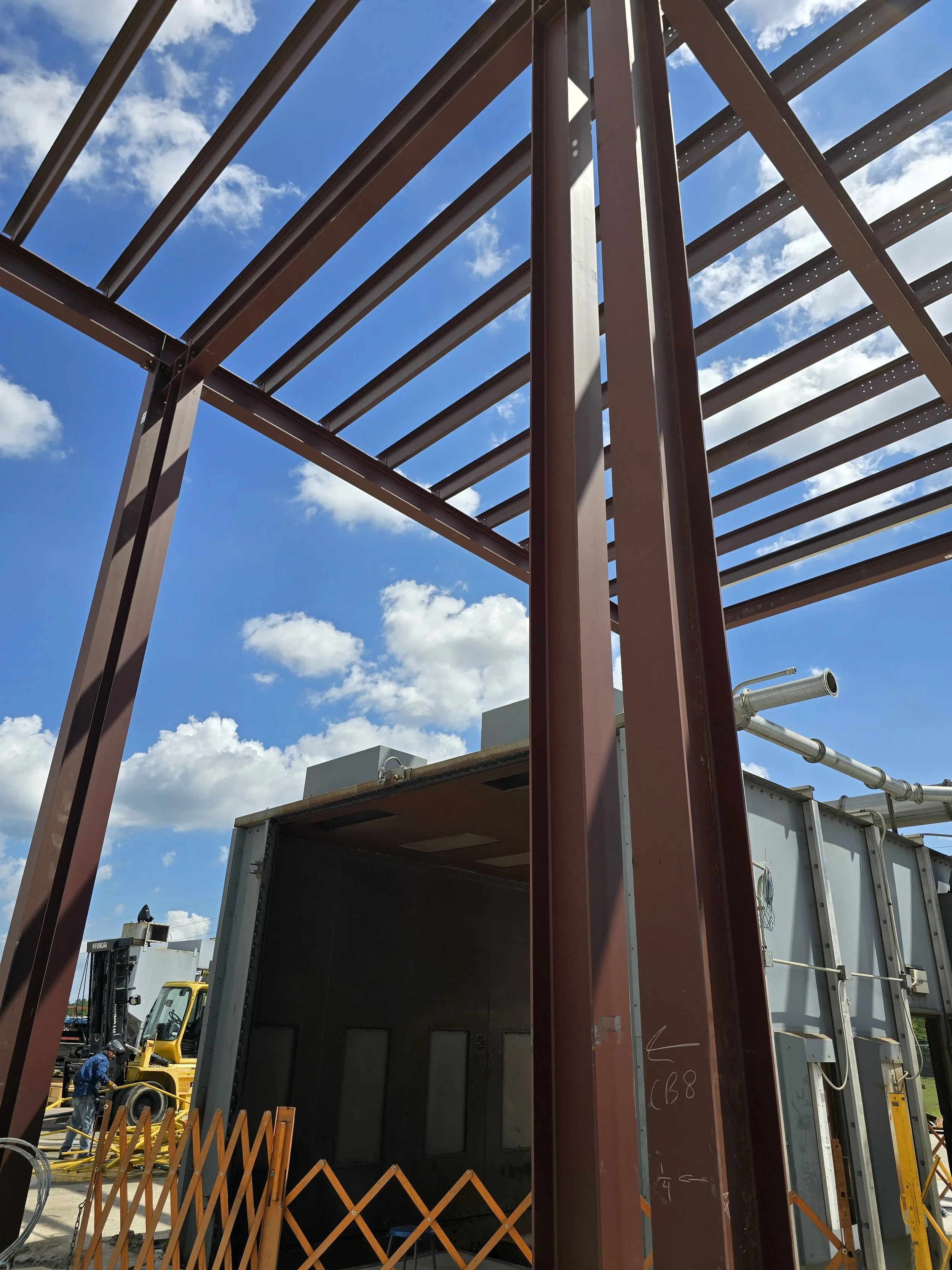 Steel framework for a building under construction, with blue sky and clouds in the background, construction workers and equipment visible at the site.