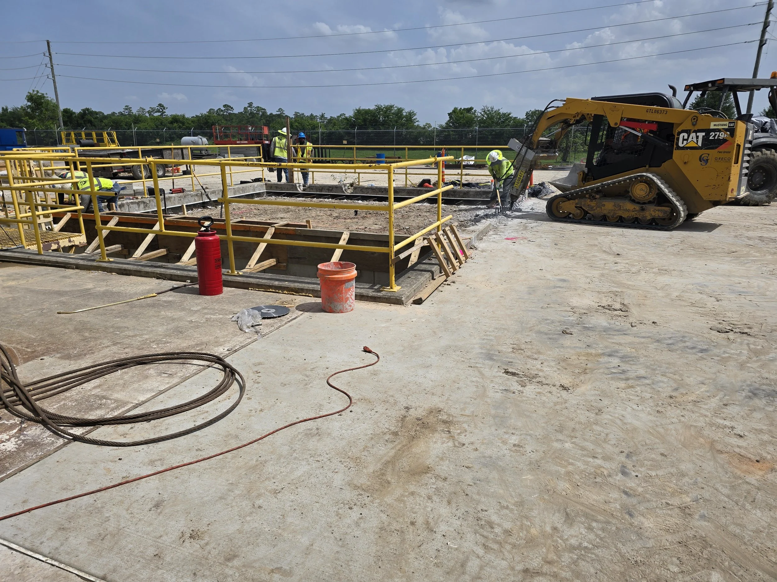 Construction site with workers, safety railings, and a small tracked excavator working on a concrete foundation.