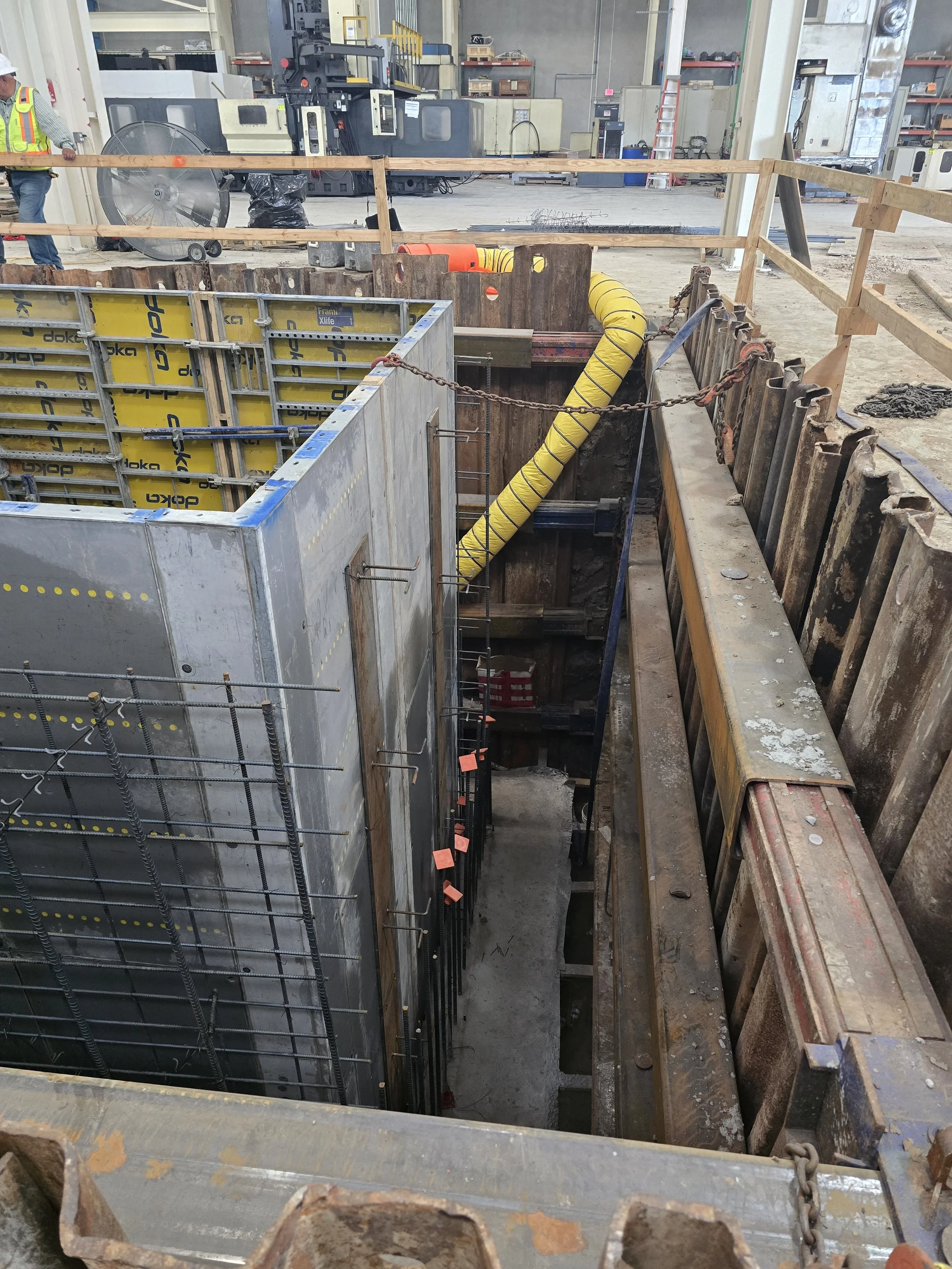 Construction site showing a deep excavated section with temporary shoring, rebar for concrete reinforcement, and workers in safety vests in the background.