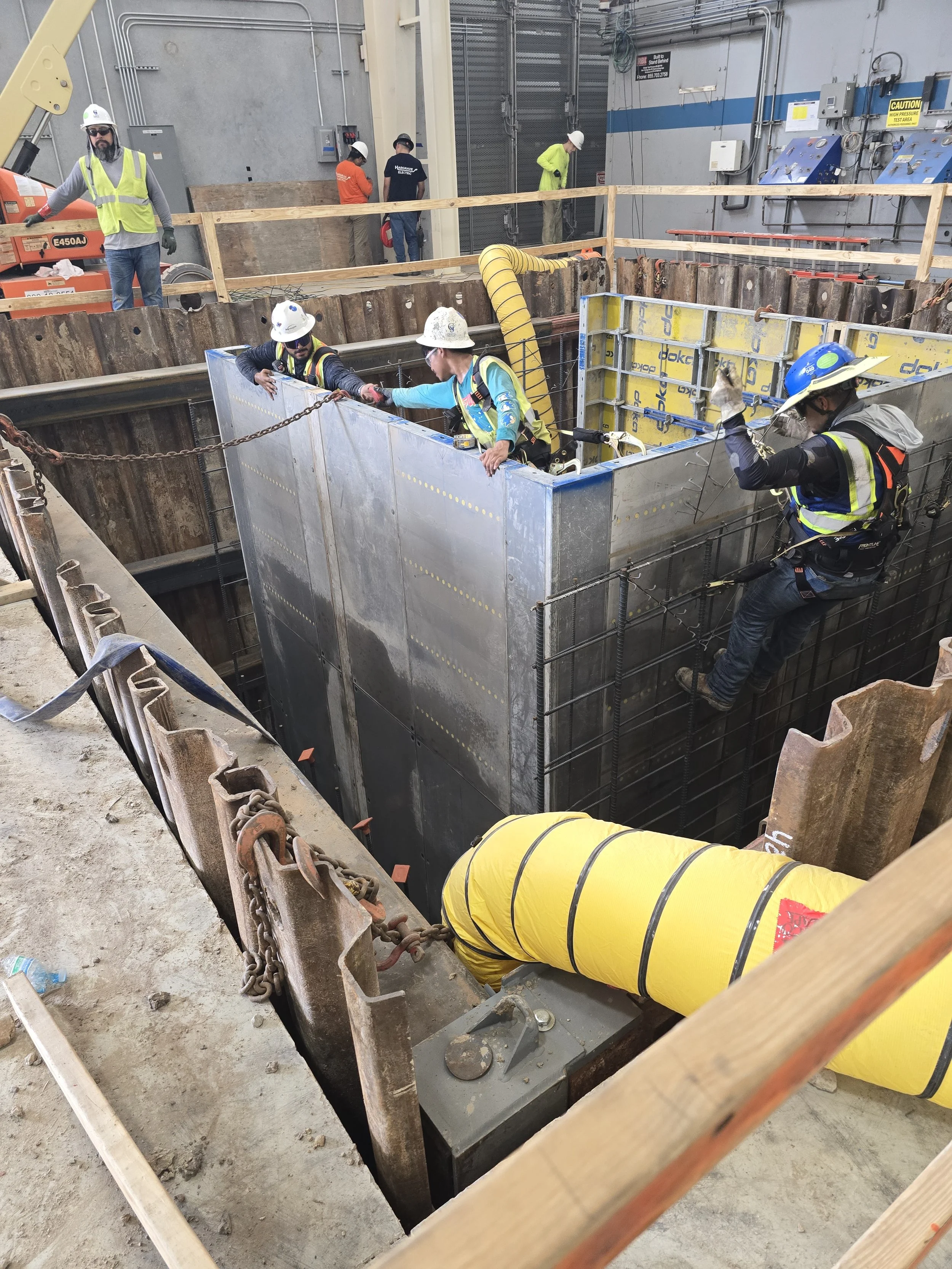 Construction workers install a large underground structure with metal panels and safety gear inside a construction site.
