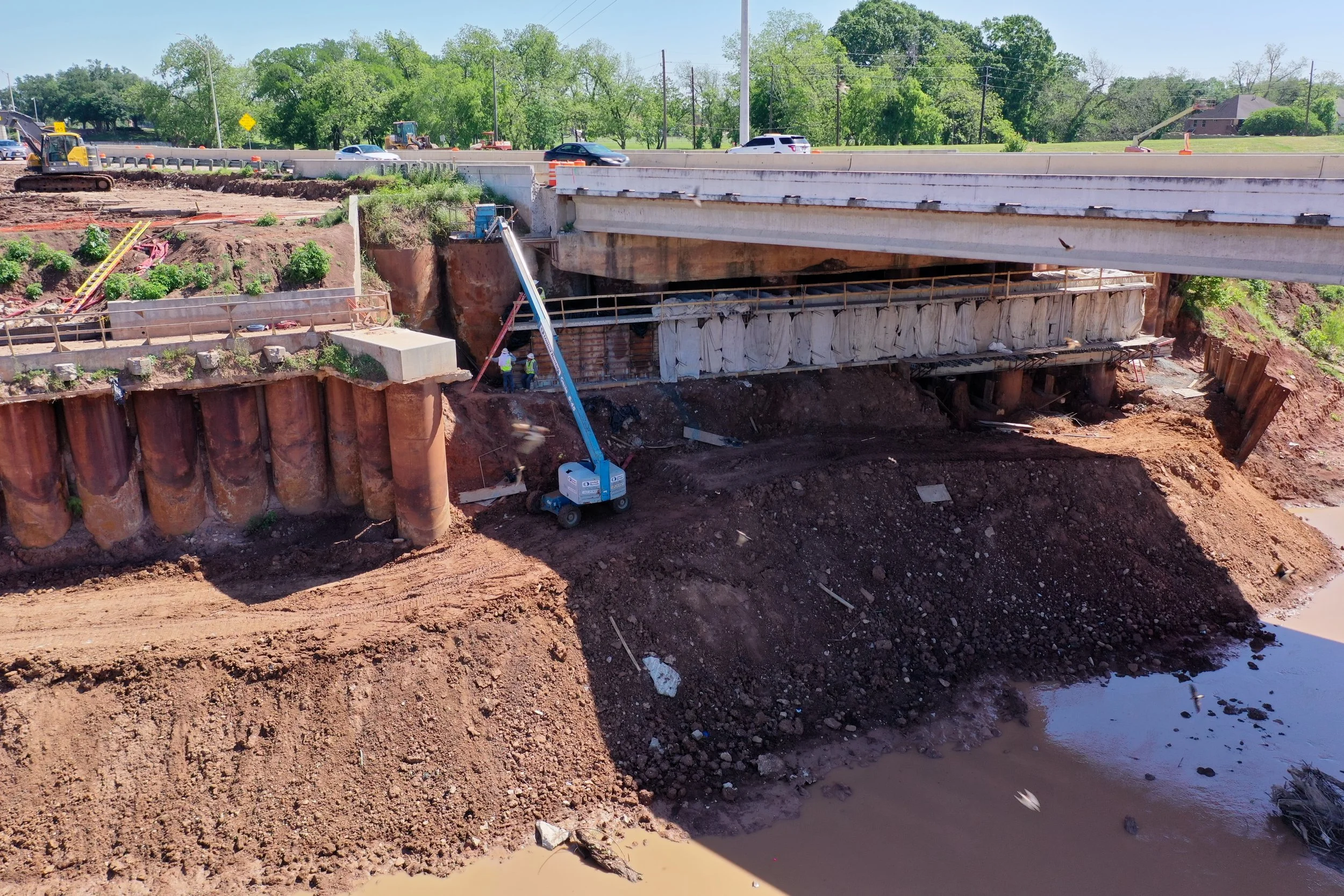 Construction workers working on a bridge repair project with heavy machinery and equipment, and exposed dirt and water underneath.