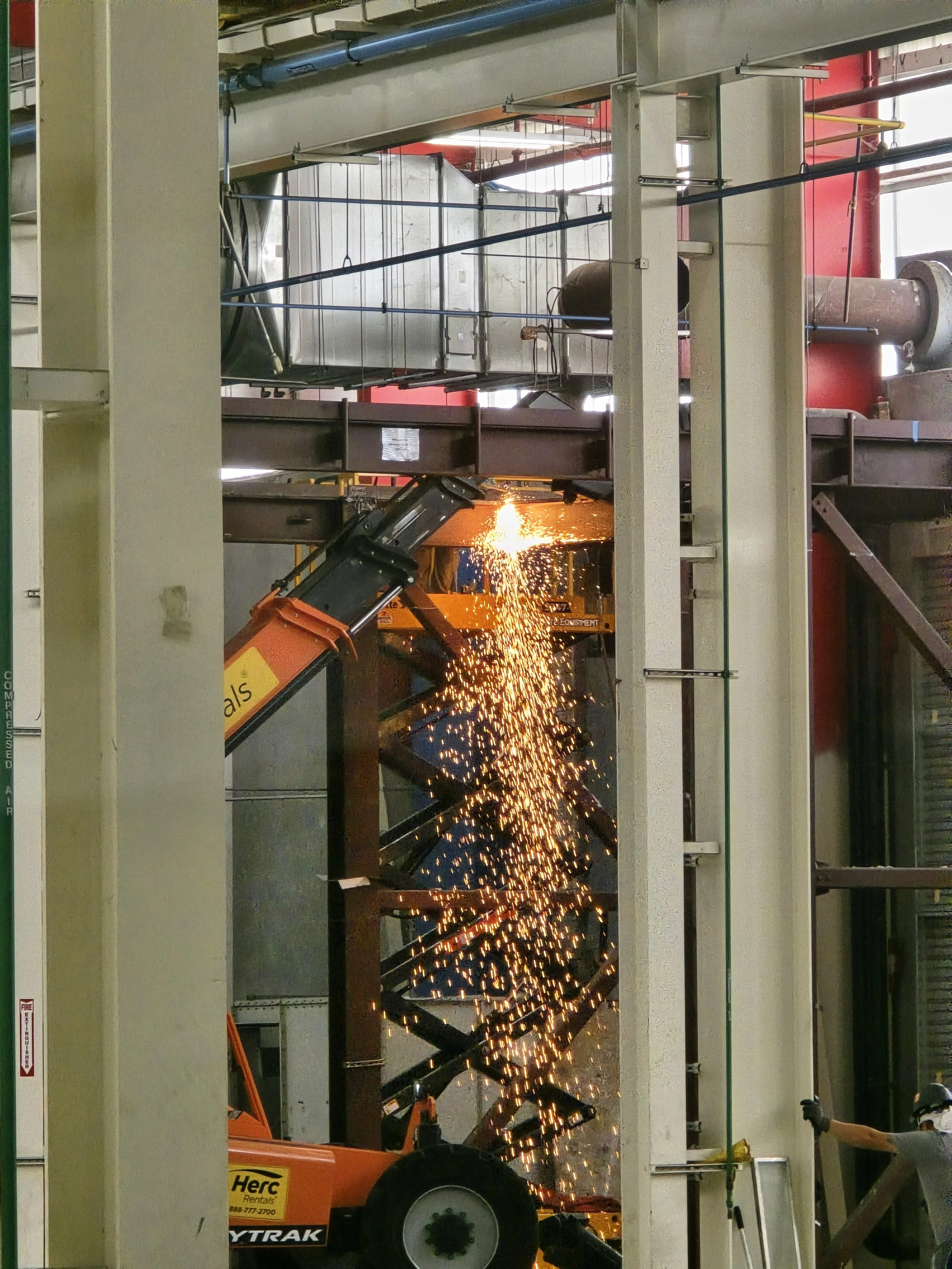 Industrial construction site with welding sparks, steel beams, pipes, and machinery, including an orange telescopic lift.