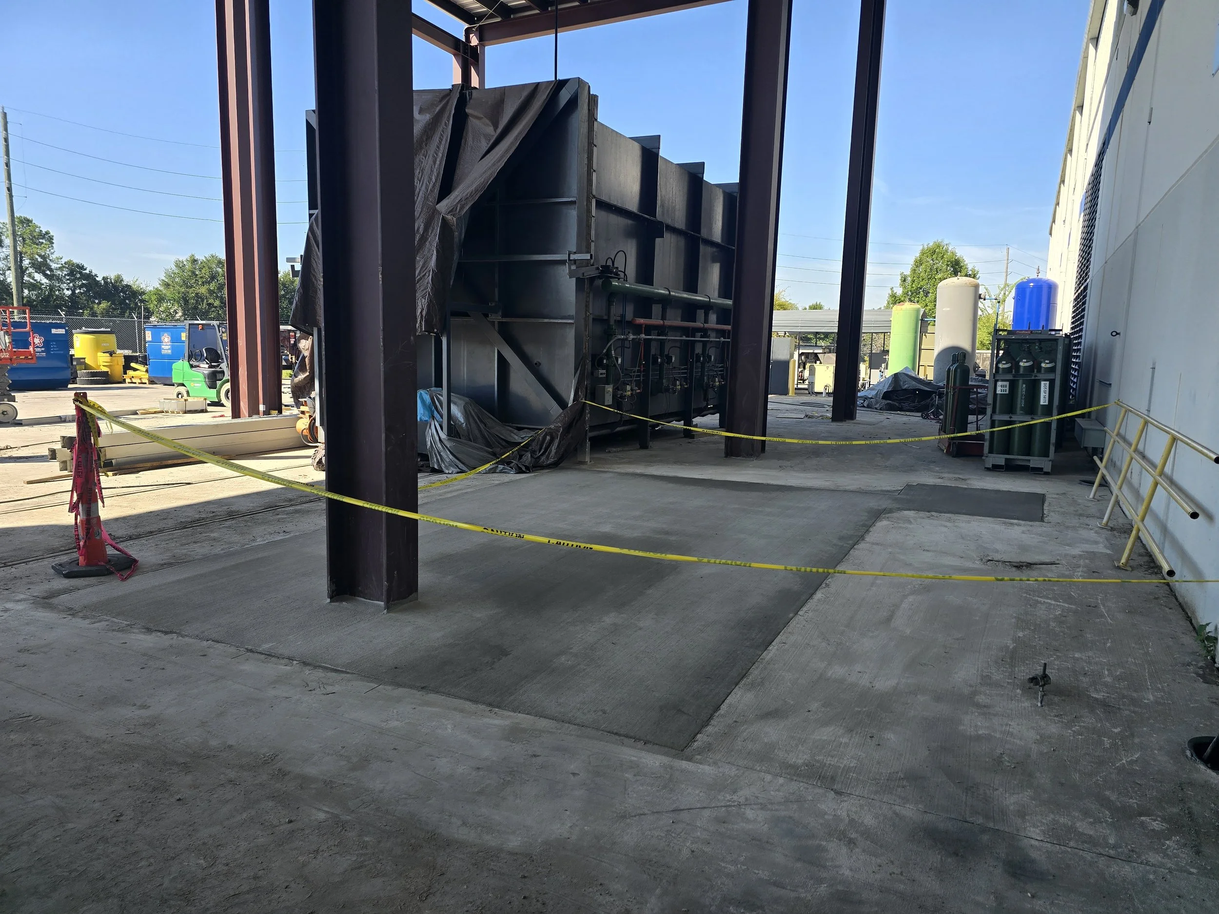 Construction area with caution tape, equipment, and structural steel framework outdoors under a clear blue sky.