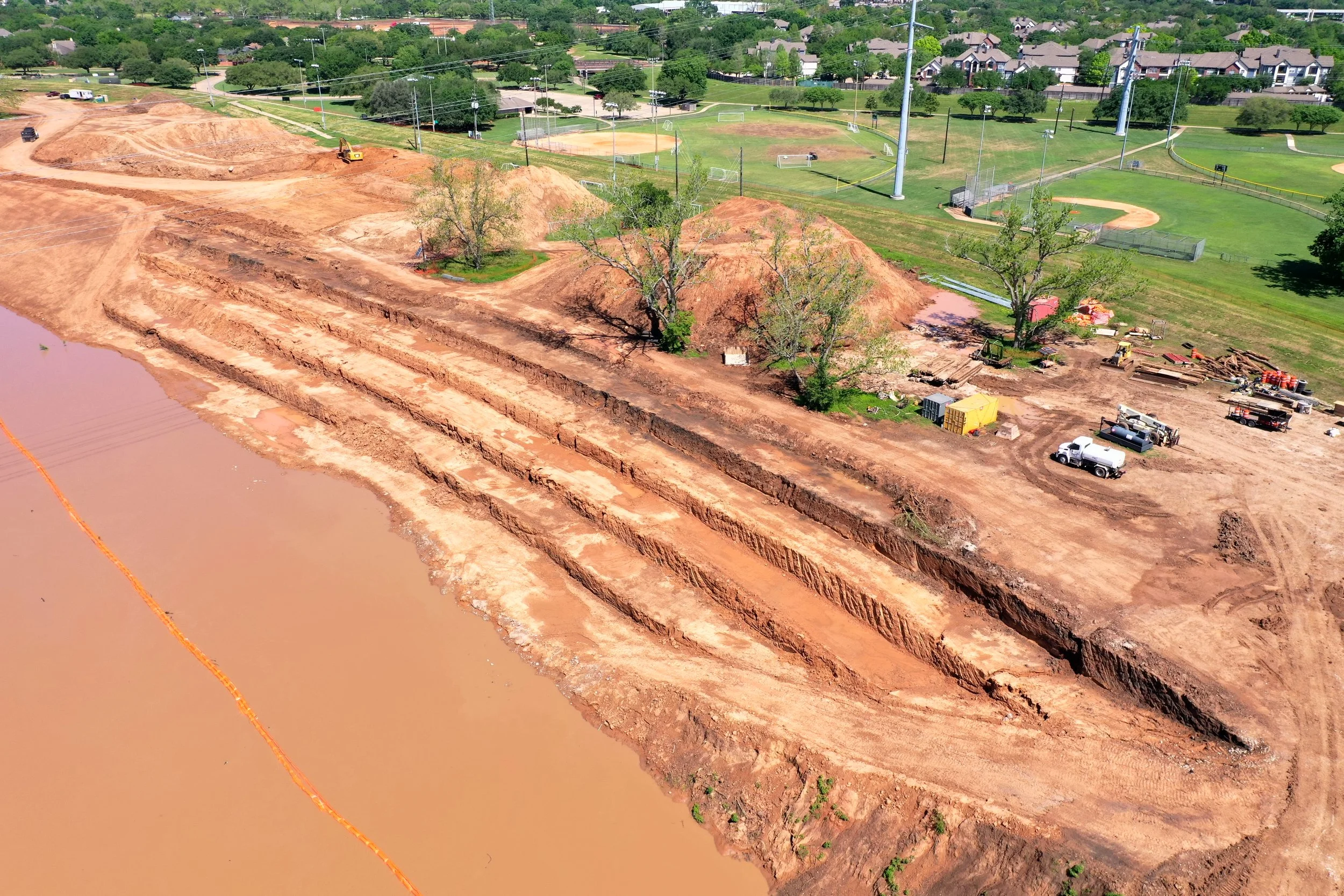 Construction site by a muddy body of water with excavated dirt and construction equipment, adjacent to a green park with sports fields.