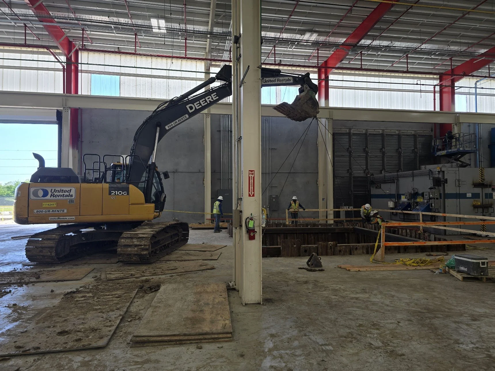 Construction site inside a large building with workers and a tracked excavator. The excavator's arm is lifting debris or material towards the ceiling, with several workers in safety gear working nearby. The area has a dirt floor with construction mat
