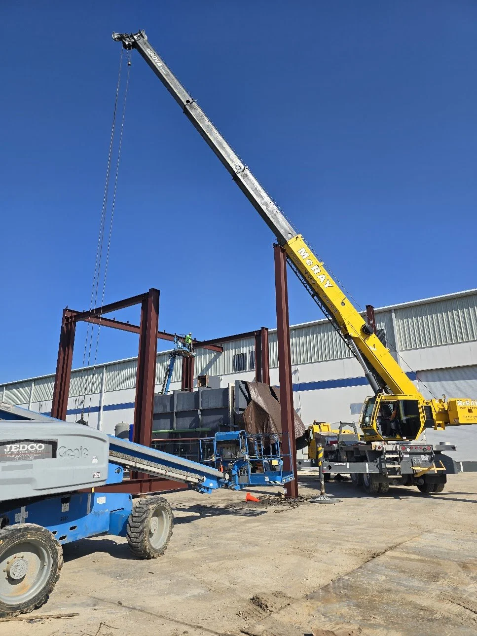 Construction site with a yellow crane lifting steel beams and a blue scissor lift on dirt ground against a clear blue sky.
