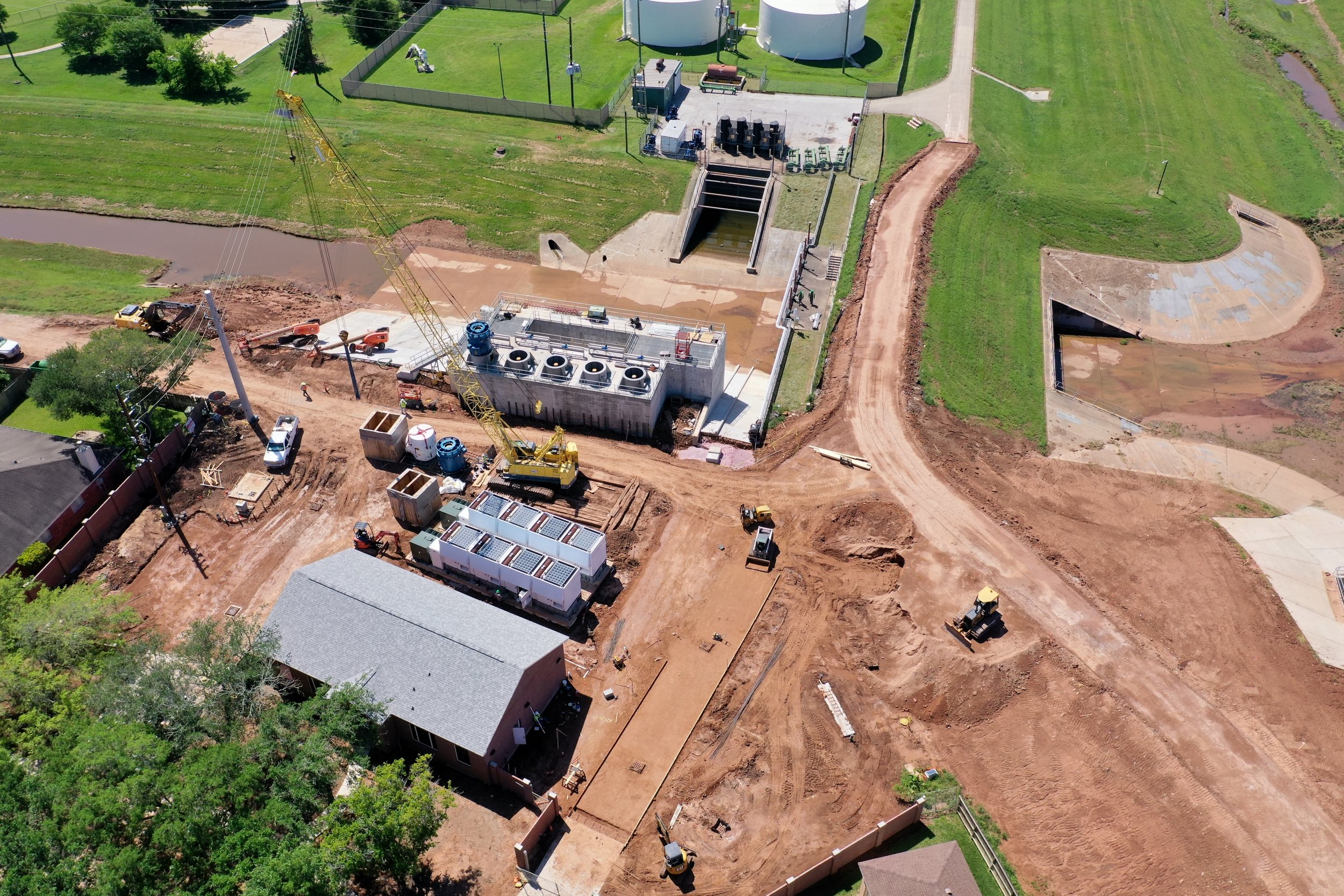 An aerial view of a construction site with heavy machinery, including a tower crane, and partially built infrastructure near residential houses and a grassy area.