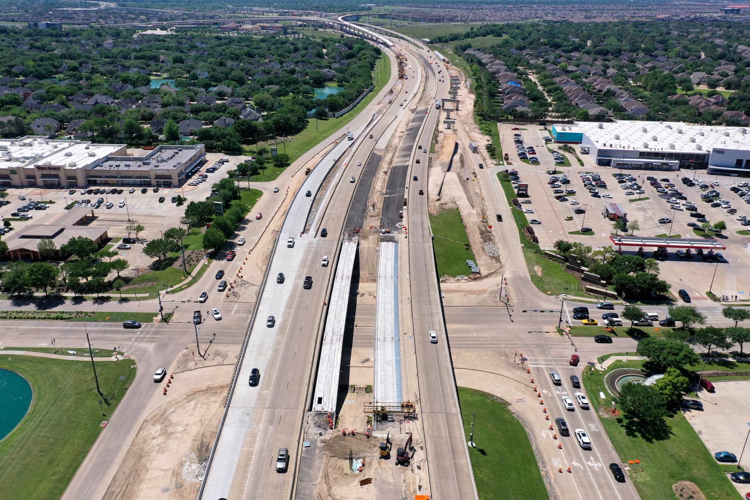 Aerial view of a highway under construction with multiple lanes, surrounded by parking lots, commercial buildings, residential neighborhoods, and green spaces.