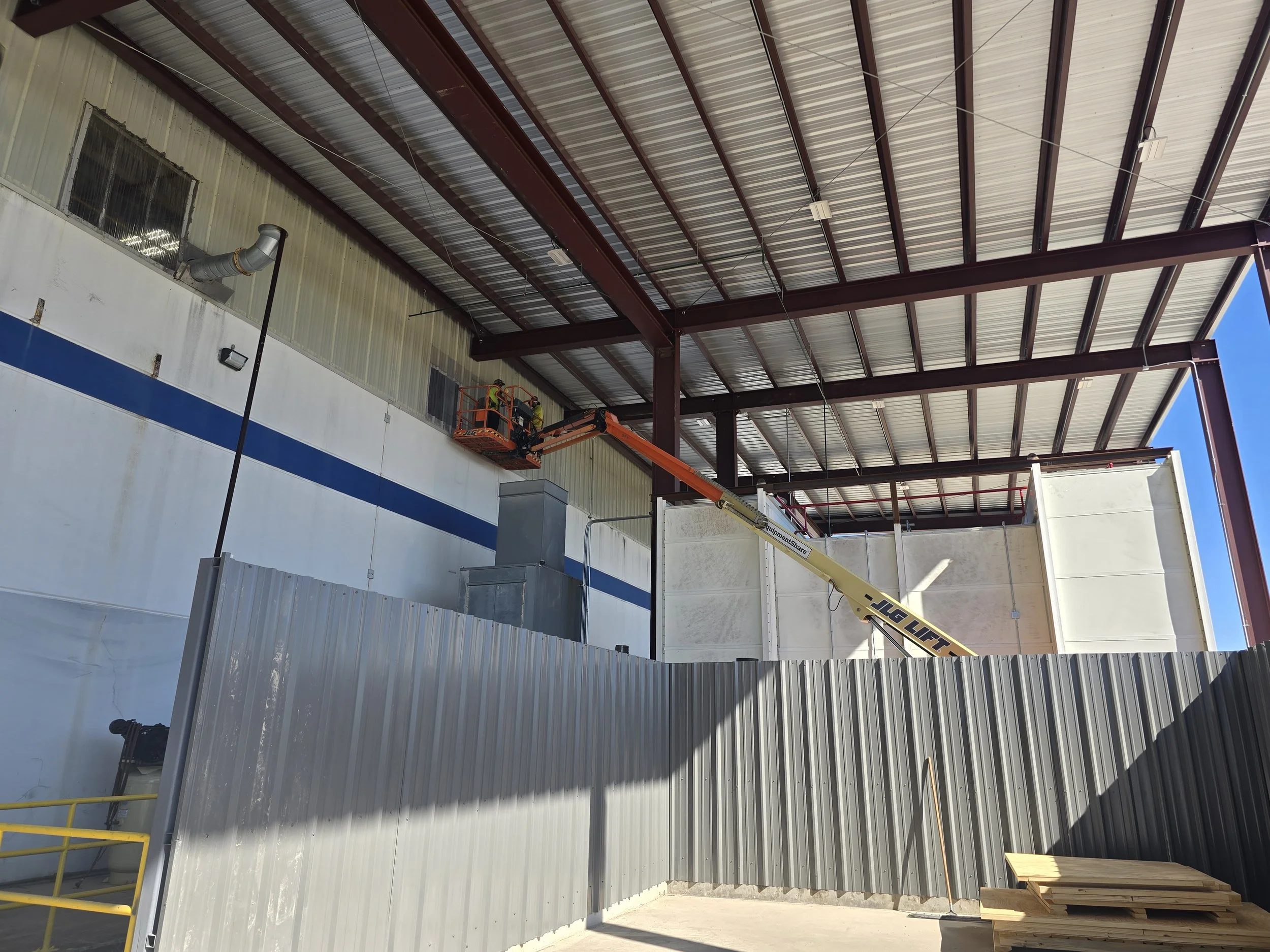 Construction workers using a lift and scissor lift to work on the wall and ceiling of a large industrial building with a metal roof and walls.