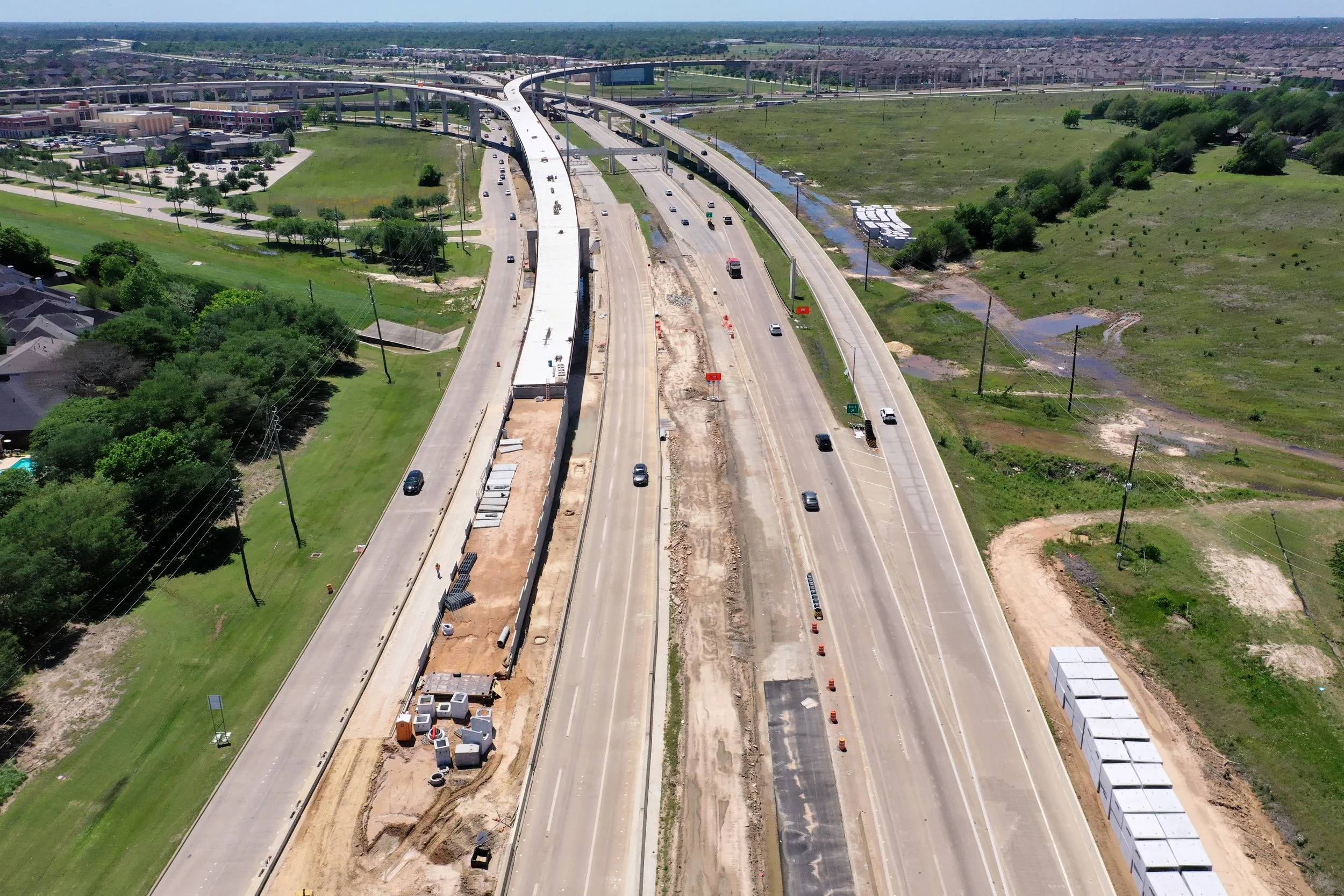 Aerial view of a highway under construction with multiple lanes, construction materials, and vehicles, surrounded by green fields and residential areas.