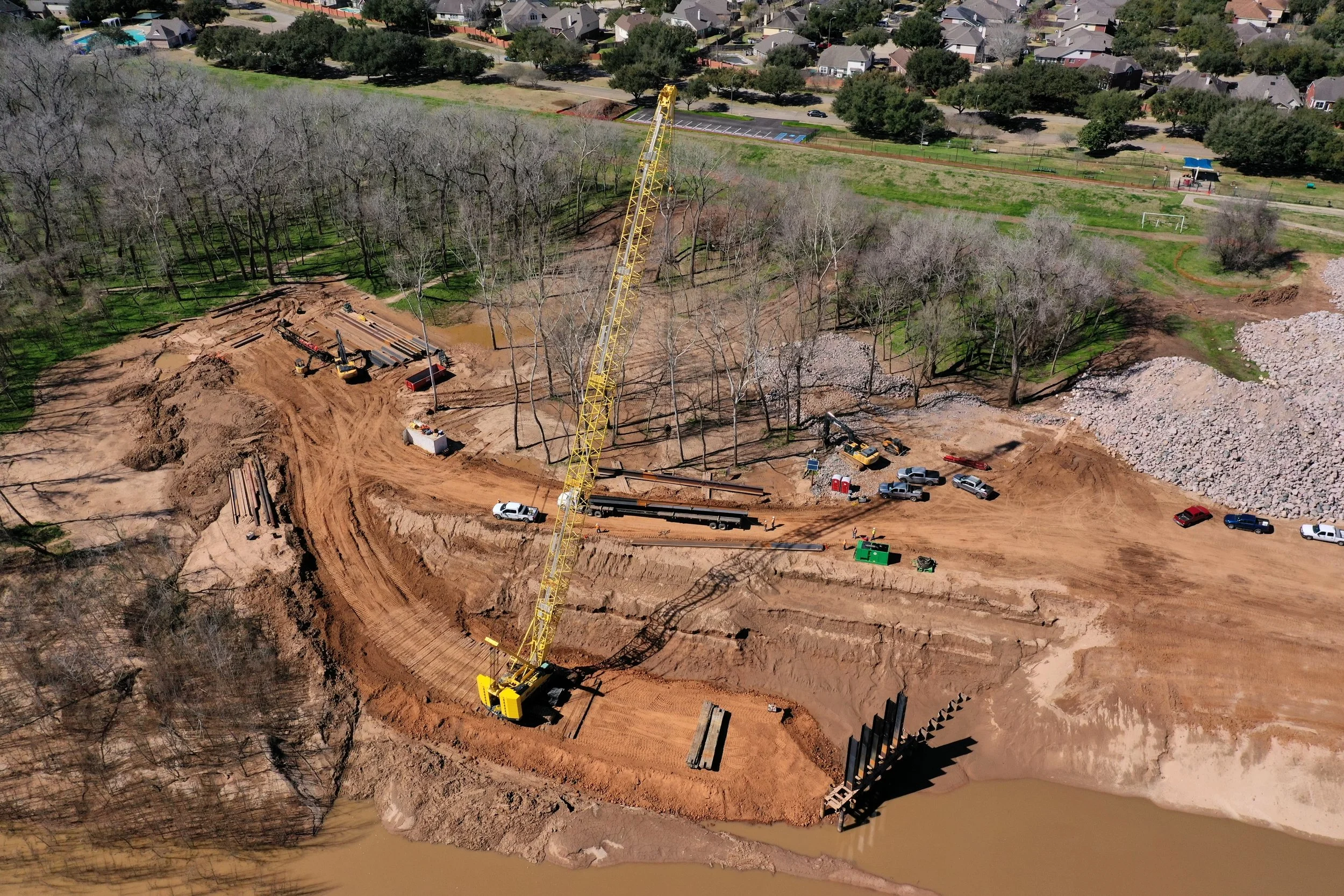 An aerial view of a construction site near a body of water, with a yellow crane, multiple construction vehicles, and dirt and gravel areas, with a wooded area and a residential neighborhood in the background.